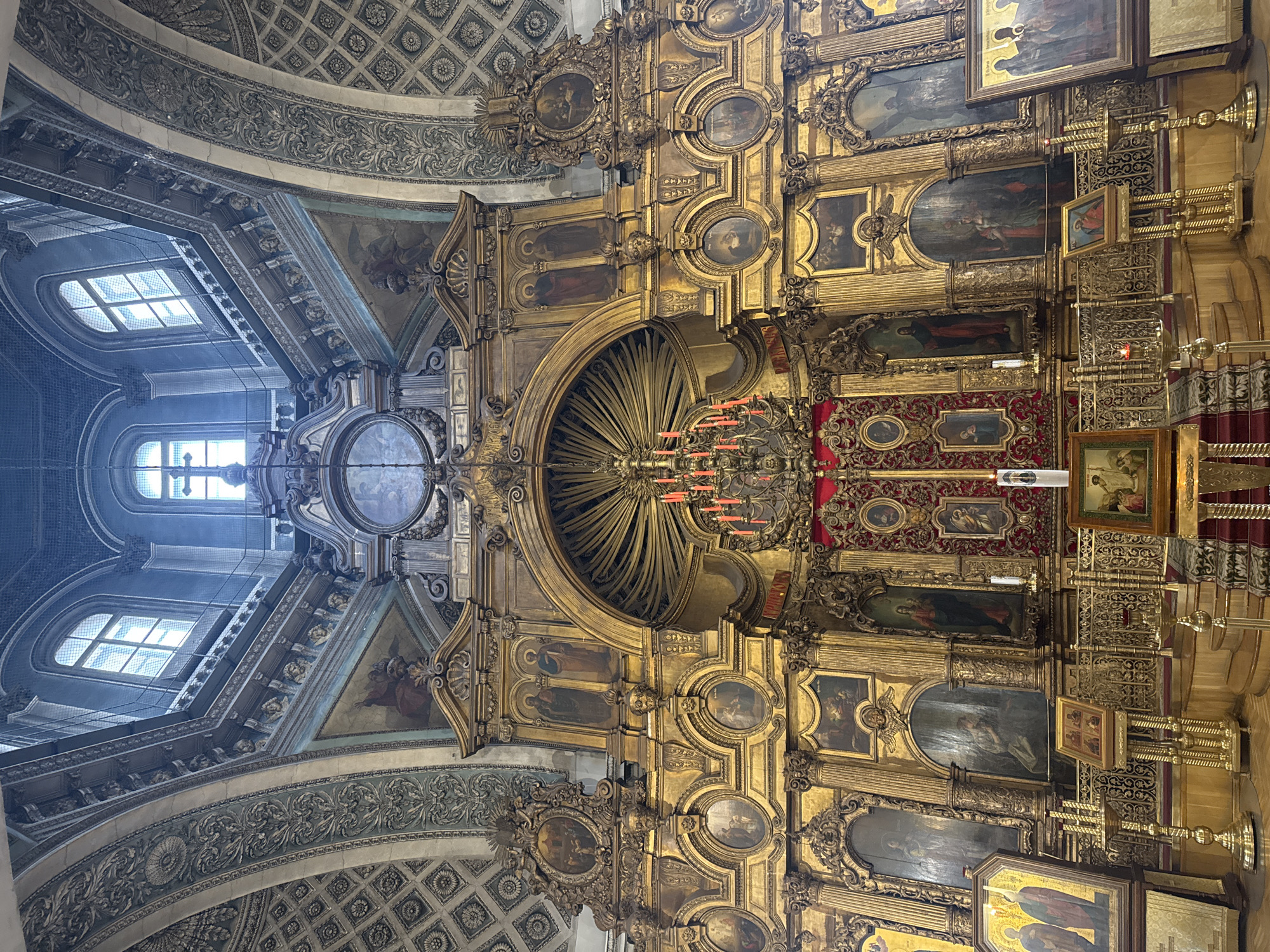 The image captures the intricate and ornate interior of a building, possibly a church or cathedral, located in Saint Petersburg, Russia. The architecture showcases heavy use of gold leafing and detailed carvings, particularly around the walls and ceilings. The lighting appears to be a mix of natural light streaming in from a high window and artificial illumination from chandeliers. Paintings and religious icons adorn the walls, further emphasizing the sacred nature of the space. The environment is grand and opulent, reflecting a rich historical and cultural context.