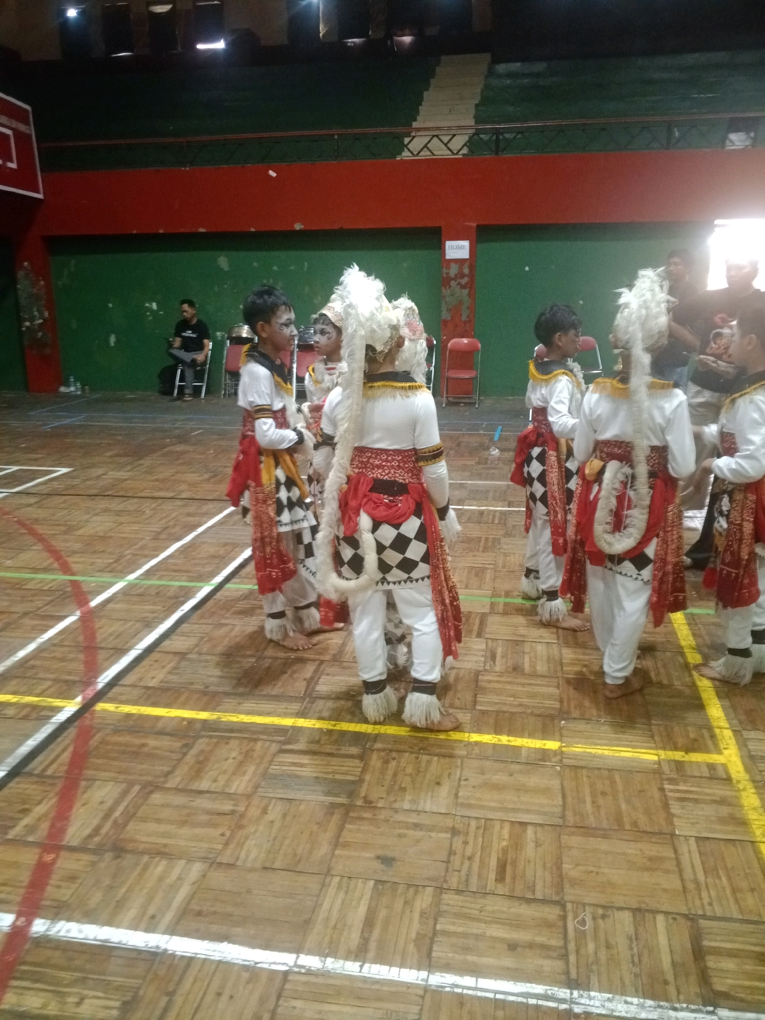 In the Wuryorejo, Indonesia, community hall, children in traditional Balinese dance attire stand on a wooden floor marked with white and yellow lines. The attire consists of white pants, elaborately decorated sashes in red and black checkered patterns, and white headdresses with feather accents. Some children have white face paint. In the background, a person is seated on a chair, and other individuals are standing. The hall features green walls and red trim, with tiered seating in the upper portion of the frame.