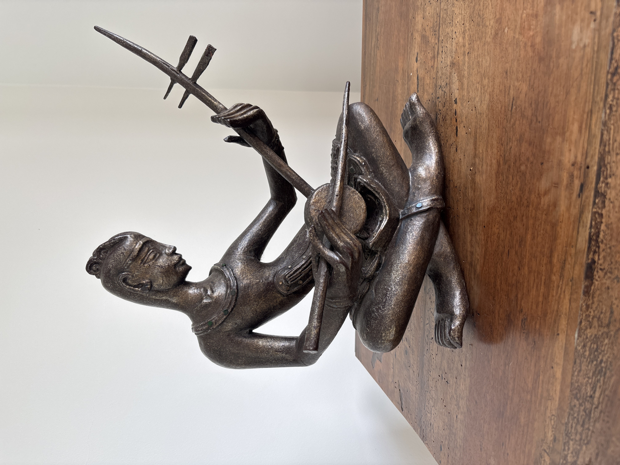 A bronze sculpture of a reclining musician is mounted on a wooden plaque against a white wall. The sculpture depicts a figure, possibly male, playing a stringed instrument. The musician's body is curved, with one arm raised to hold the neck of the instrument and the other hand positioned near the sound box. The instrument has tuning pegs along the neck. The figure has a small topknot on the back of the head, and wears a simple piece of jewelry. The wooden plaque is a rectangle with visible wood grain. The background is a plain white wall. There are no specific visual indicators to confirm the location as Ban Bang Khao, Thailand, although that may be where this type of art is common.