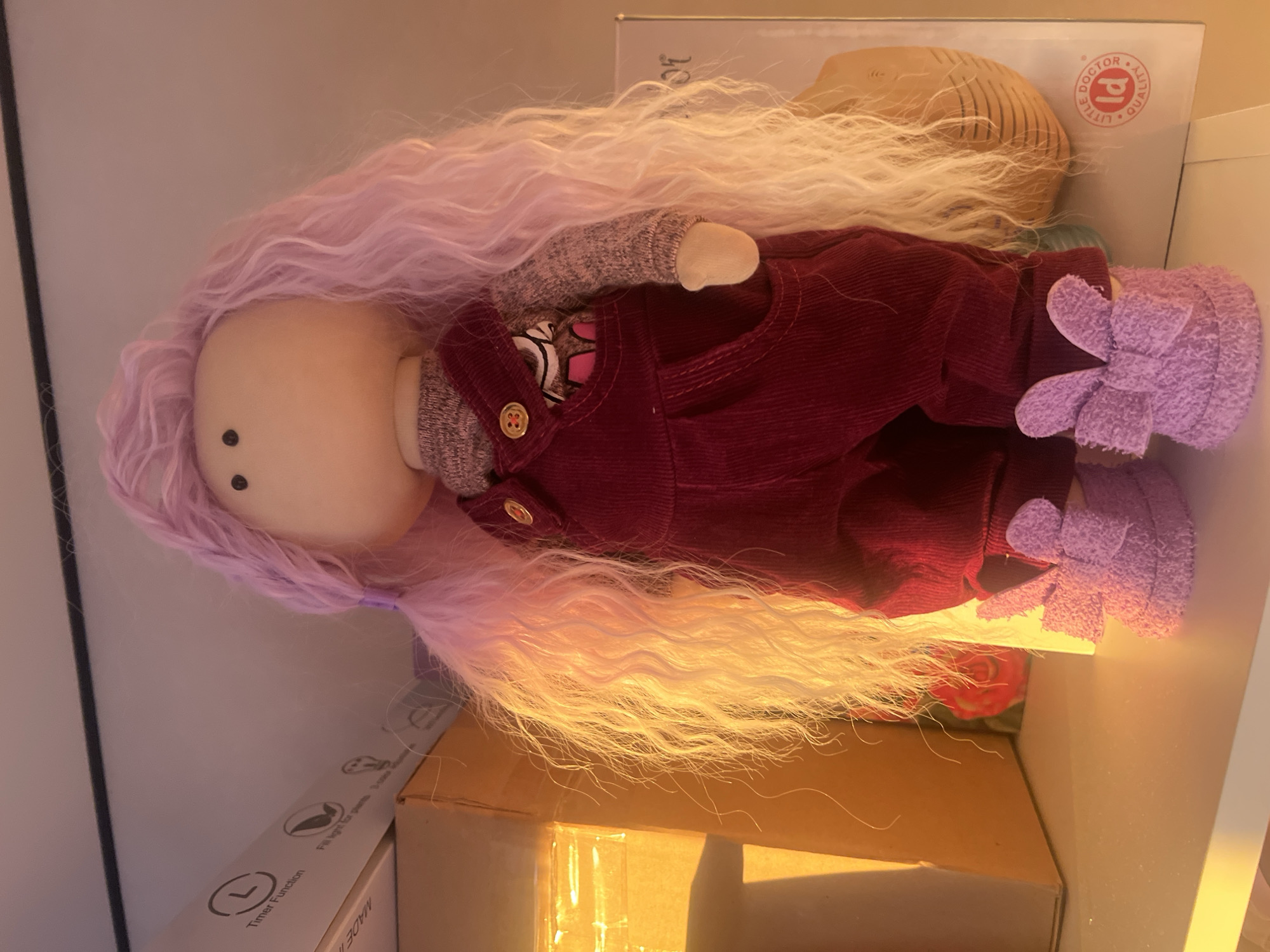 A cloth doll with light purple hair, maroon corduroy overalls, a gray shirt, and purple flower-decorated shoes is lying on its side. The doll has two black eyes but no other facial features. The doll is lying on top of cardboard boxes. One box has white text and logos on it. Another box has "LITTLE DOCTOR QUALITY" printed on its surface. The scene appears to be indoors. There is no indication that this image was taken in Votkinsk, Russia.