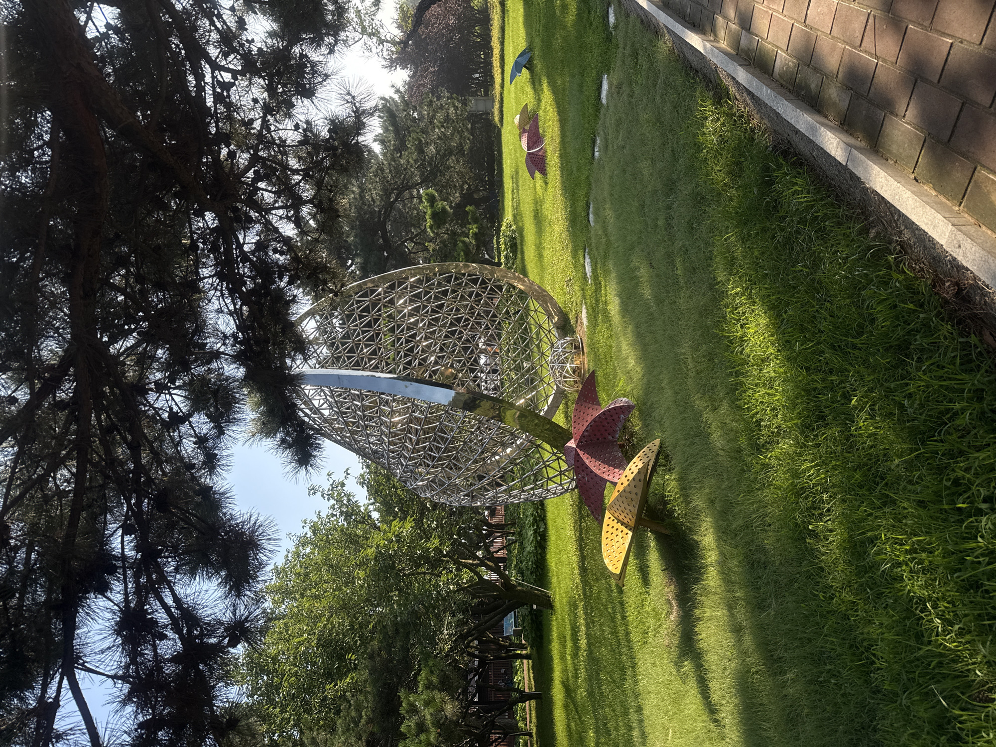 In the city of Qinhuangdao, China, a park setting features a large, spherical metal sculpture composed of a intricate geometric lattice. In the foreground, three abstract, mushroom-like sculptures are visible on a manicured green lawn. The largest sculpture, in yellow and red hues, is closer to the viewer. Further back, two smaller, umbrella-shaped sculptures, one pink and one blue, rest on the grass. The scene is framed by the branches of a large pine tree overhead. A paved walkway borders the lawn on the right side of the image.