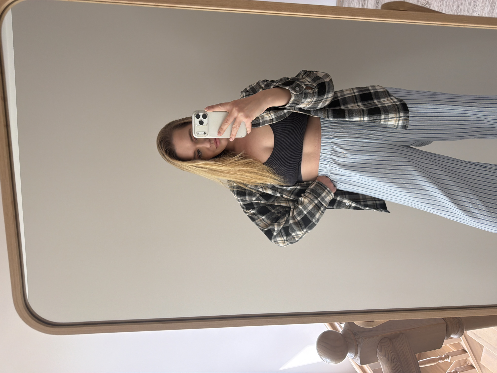 A person with long blonde hair takes a selfie in a mirror, holding a white smartphone. The person wears a black sports bra, a plaid shirt, and blue and white striped pajama pants. The background shows a neutral-colored wall and parts of a wooden frame and fixture attached to the mirror. The location appears to be indoors, not explicitly identifiable as Dreiliņi, Latvia.