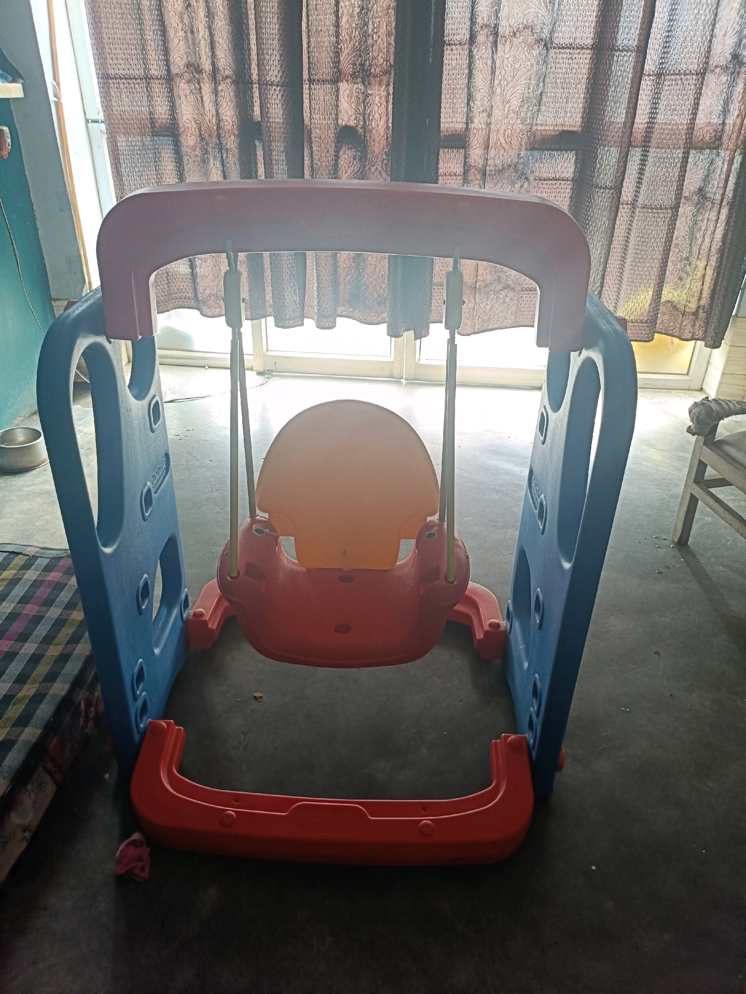 The image shows a children's plastic swing set, primarily red and blue, positioned indoors. The swing set features a red seat suspended from a red-and-blue frame. The background consists of patterned brown curtains covering a window or door, and a light-colored floor. To the left, a blue plastic object, possibly a part of another toy or furniture, is visible along with a patterned cloth. A metal bowl is also present to the left of the swing set. The overall scene appears to be in a residential setting, likely a home in Mohanlalganj, India, given the context. There are no people visible in the image.