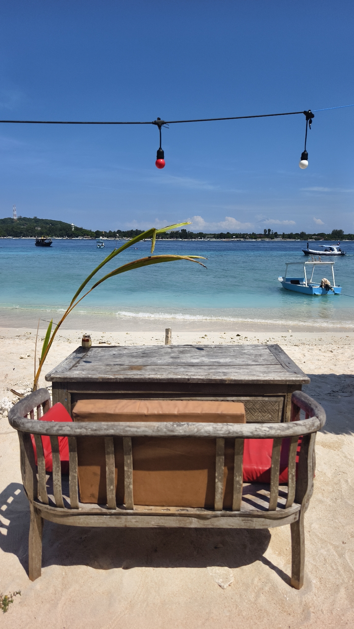 The image depicts a beach scene at an outdoor seating area in the city of Unknown, Indonesia. In the foreground is a weathered wooden bench with red cushions and a brown backrest. A wooden table is positioned behind the bench, with a thin, green plant leaning against it. Beyond the seating area is a white sandy beach meeting clear, turquoise water. Two small boats are anchored in the water. The background shows a hazy island with green vegetation and what appears to be a radio tower. Above the seating area, string lights with red and white bulbs are suspended. The sky is a clear, bright blue. The lighting suggests a sunny day. The scene appears tranquil and tropical.