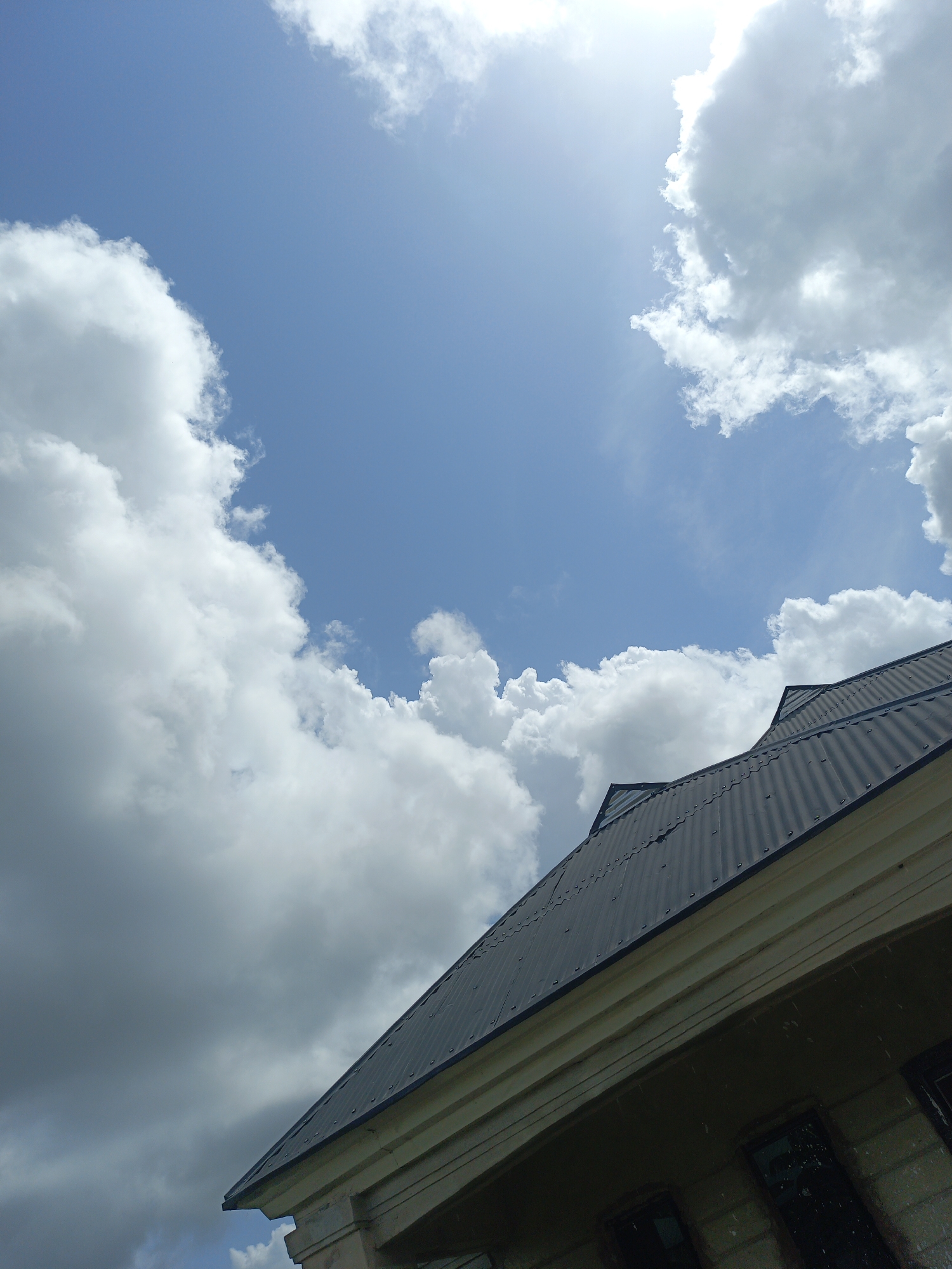 The image shows a building with a dark, corrugated metal roof, viewed from a low angle. The roofline angles upwards from the lower right to the upper left of the frame. Below the roof, a light-colored structure with what appears to be windows can be seen. Above the building, the sky is visible, with patches of blue sky interspersed with large, white, puffy clouds. The location is listed as Unknown, Unknown. There are no people visible in the image.