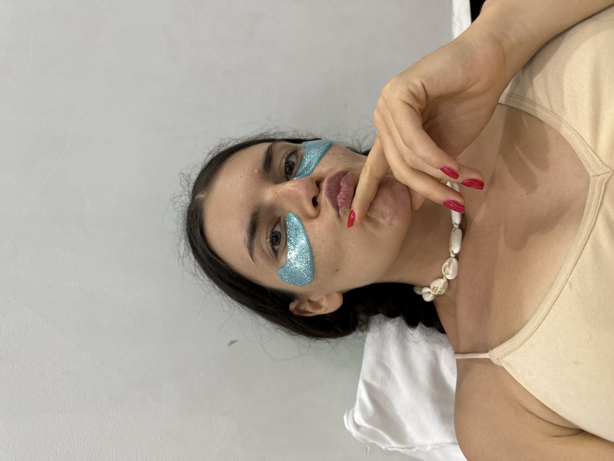 A young woman with dark hair pulled into a braid and adorned with blue glittery under-eye patches is resting on a white pillow in Mũi Né, Vietnam. She wears a light beige camisole and a necklace made of white shells. Her hand is raised to her chin, with bright red nail polish visible on her fingernails. The background is a plain, textured, light-colored wall.