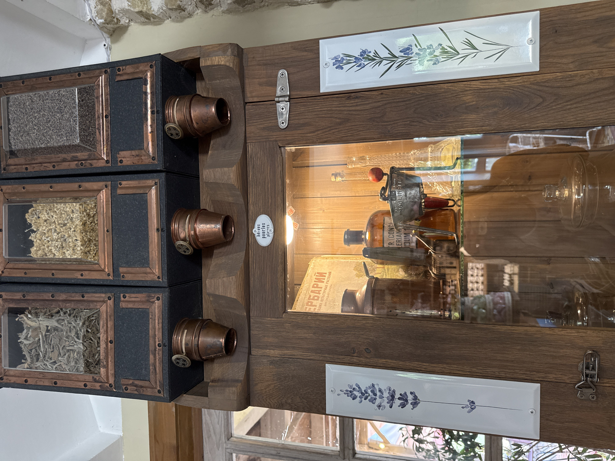 In Semogorskiy, Russia, a vintage wooden apothecary cabinet stands against a stone wall. The upper section features three glass-fronted compartments filled with dried herbs and seeds, each with a copper dispensing mechanism below. The main body of the cabinet is a wooden case with a glass door, revealing shelves stocked with antique medicinal bottles, a copper jug, a tin labeled "MOULINETTE," and other assorted glass containers. Two decorative ceramic tiles, one on each side of the cabinet door, depict sprigs of lavender. The lighting suggests an interior setting, possibly a shop or a historical display.