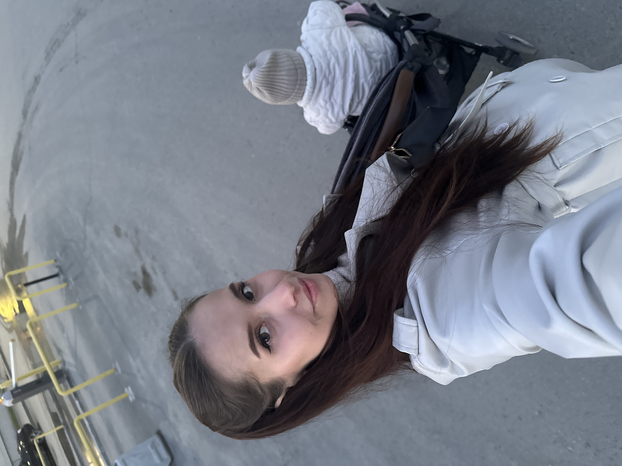 The image captures a high-angle, selfie-style view of a young woman and a baby outdoors, likely in Yekaterinburg, Russia. The woman, positioned in the foreground, has long dark brown hair and is wearing a light-colored (possibly off-white or light grey) trench coat or jacket over a turtleneck. She looks directly at the camera with a neutral to slightly pleasant expression.
Behind her, partially visible, is a baby or small child seated in a dark-colored stroller with brown accents. The child is dressed in a light beige knitted hat and a white quilted jacket. The ground beneath them is a paved surface, resembling concrete or asphalt. To the left, several yellow metal barriers or railings are visible, suggesting a public area such as a pedestrian zone or park.
The lighting is soft and diffused, indicating an overcast day or a time of day with indirect sunlight, like early morning or late afternoon. No harsh shadows are present. The scene suggests a casual outdoor activity, such as a walk with the baby. There is no discernible text within the image.