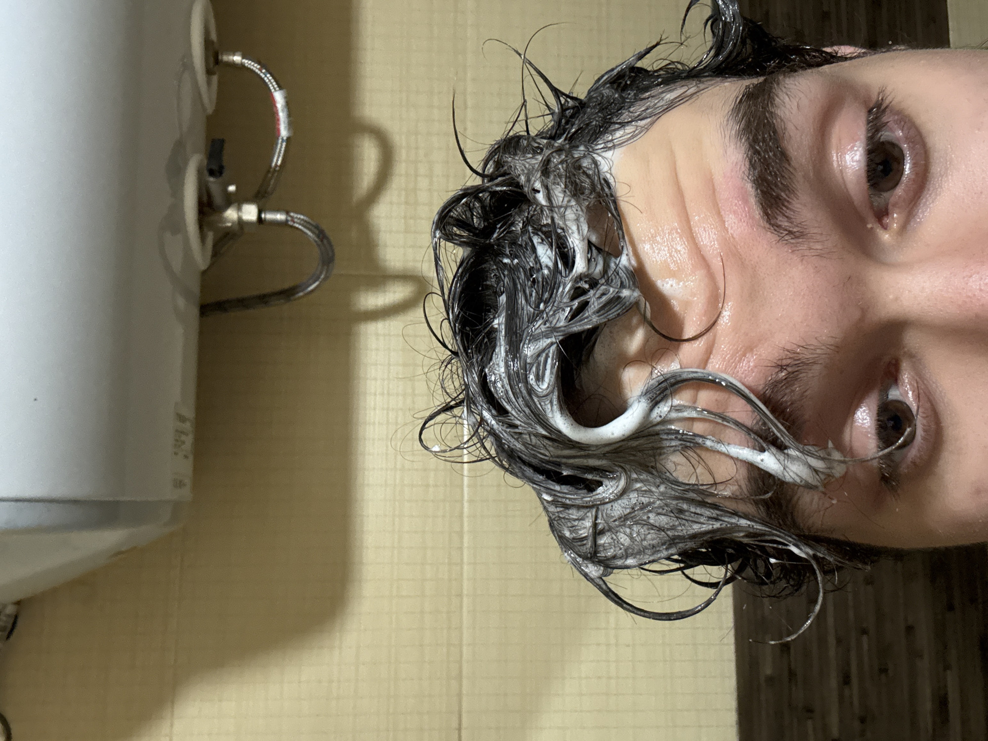 A close-up, low-angle indoor photograph depicts the upper portion of a person's head and an overhead appliance, located in Blagoevgrad, Bulgaria. The subject, an adult with dark hair, is shown actively washing their hair, which is coated in white lather. The eyes are visible, looking directly towards the viewer or slightly downward. The forehead exhibits minor lines. Above the subject's head, a white, rectangular electric water heater (boiler) is mounted horizontally on a beige tiled wall. Two flexible metallic hoses with associated fittings extend from the underside of the water heater, one with a visible red marking. The wall behind the subject and the appliance consists of light-colored rectangular tiles. The bottom edge of the frame reveals a darker, textured surface, possibly a different type of tile or paneling. The scene is illuminated by artificial light.
