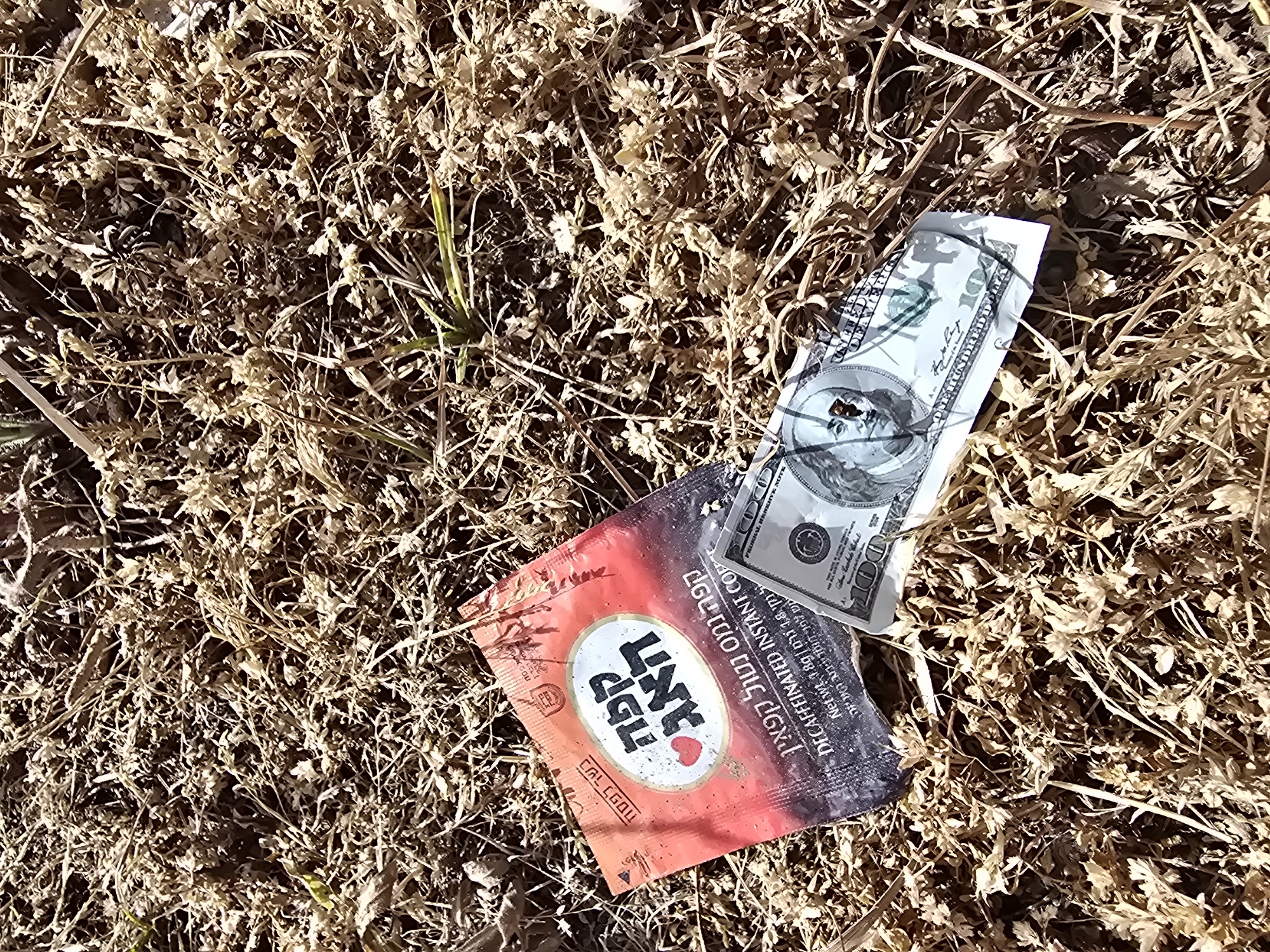 The image shows litter on the ground, consisting of a crumpled package of decaffeinated instant coffee (Hebrew text visible) and a partially visible US $100 bill held together with a thin wire. The background is dry, brown vegetation. The location is not verifiable from the image alone.