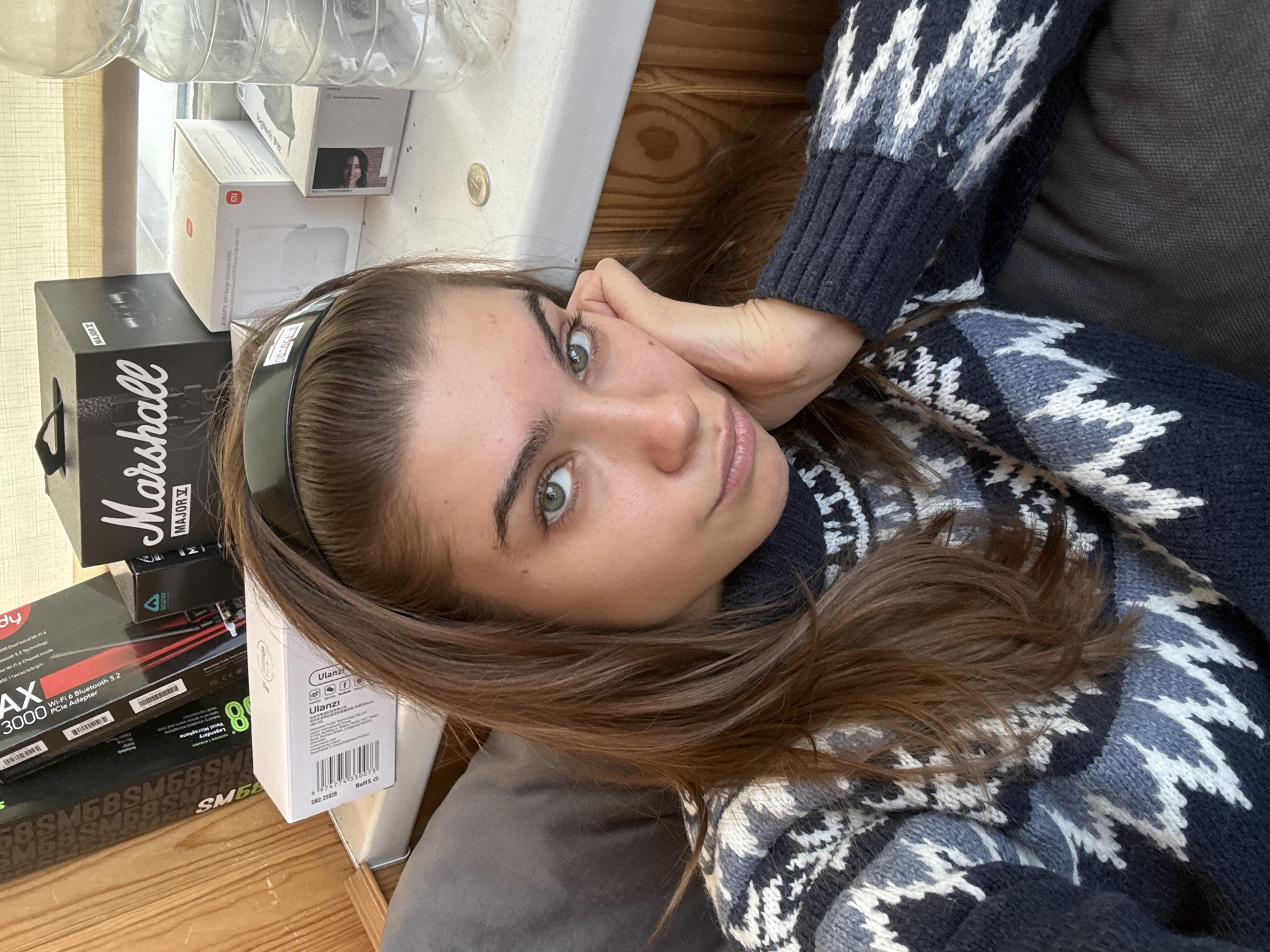 A woman with brown hair and green eyes wearing a patterned navy and white sweater is lying down, her head resting on a pillow. She is wearing a black headband. Her right hand is touching her cheek. Several boxes are stacked to her left, including boxes for a Marshall Major I headset, a TP-Link Archer TX3000 WiFi card, a Ulanzi product, a Shure SM58 microphone, and a Xiaomi WiFi range extender. A plastic water bottle is visible above the boxes. The scene appears to be indoors. No information about the location in the city of Khimki, Russia can be obtained from this image.