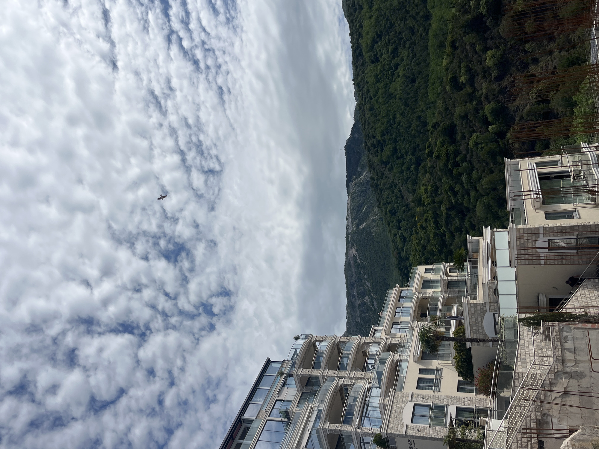 The scene depicts Bečići, Montenegro on a cloudy day. A multi-story building with balconies is located on the right. Behind the building, a mountain covered in green trees is visible. A helicopter is flying in the cloudy sky. Building construction is present in the right portion of the image.