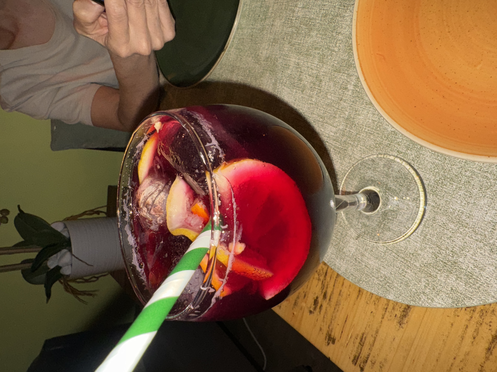 A glass of Sangria with a green and white striped straw sits on a table with a green placemat. An orange plate is to the right of the glass. A green plate and a person's arm are to the left. The table is wood. In the lower left corner is a small plant in a white pot. The drink contains red wine and sliced fruit. The location is Barcelona, Spain.