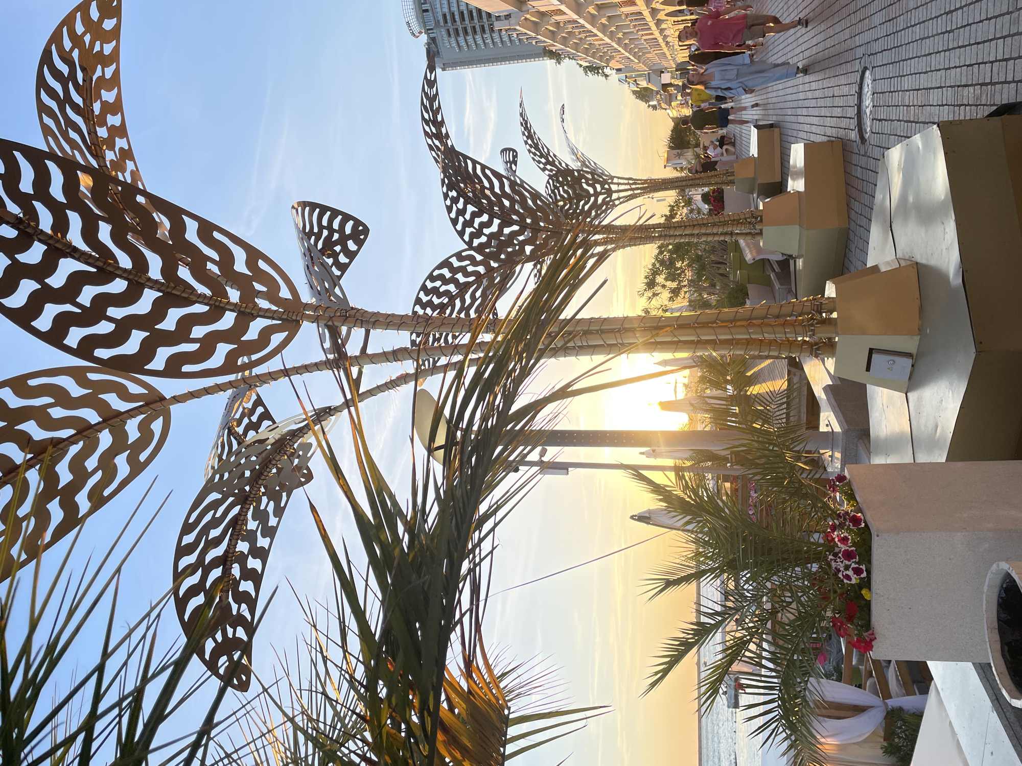 This image depicts a lively street scene in Sochi, Russia, at sunset. The foreground is dominated by ornate, metallic palm tree sculptures with intricate leaf patterns, flanking a paved walkway. To the left, lush green palm fronds partially obscure the view, with a potted plant filled with vibrant red and white flowers in the immediate foreground.
Beyond the sculptures, the walkway stretches into the distance, lined with people strolling and enjoying the evening. The architecture on the right suggests a row of buildings, possibly hotels or shops, with balconies and awnings. The setting sun casts a warm glow over the scene, illuminating the sky with golden hues and reflecting off the water in the distance, indicating the proximity to the Black Sea.
In the background, a tall, modern building stands out against the horizon. The overall atmosphere is one of a popular seaside resort, bustling with activity as day transitions into evening. The presence of decorative elements like the sculptures and potted plants suggests a well-maintained public space.
