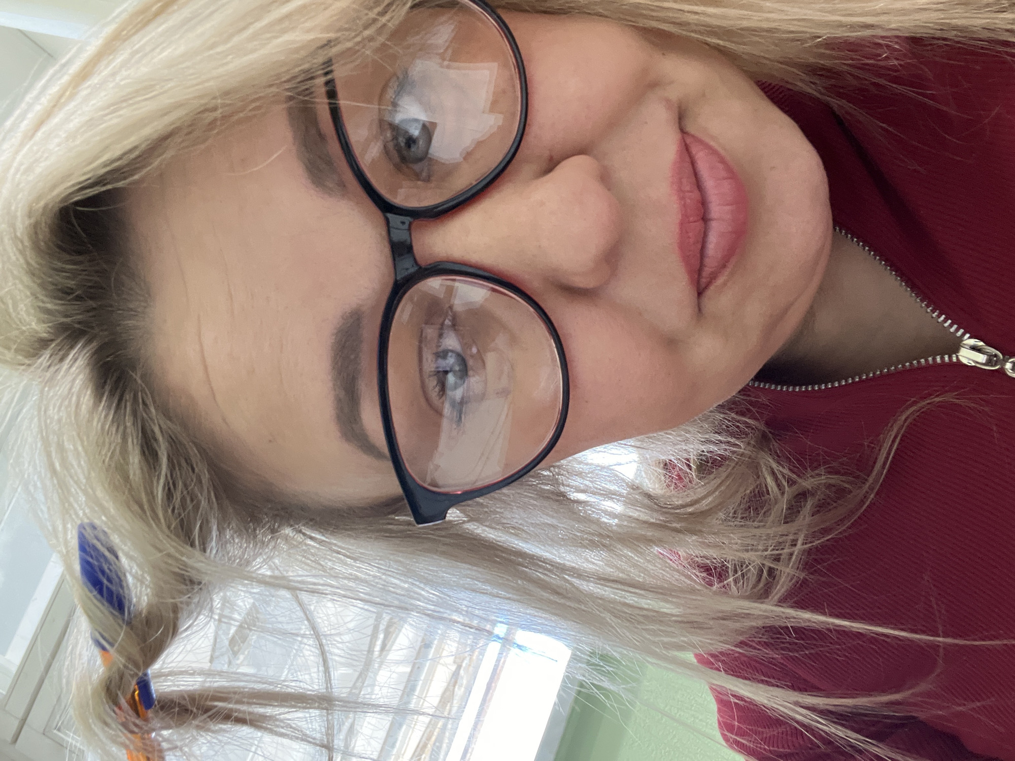 A close-up, inverted selfie photograph depicts a fair-skinned woman with blonde hair and blue eyes wearing round glasses with a black frame. Light is reflecting off the lenses. The woman is wearing a red top with a visible zipper. A blue and orange hair accessory is in her hair. The background suggests a window with a light-colored frame and possibly a green wall. Location information cannot be deduced from the image.