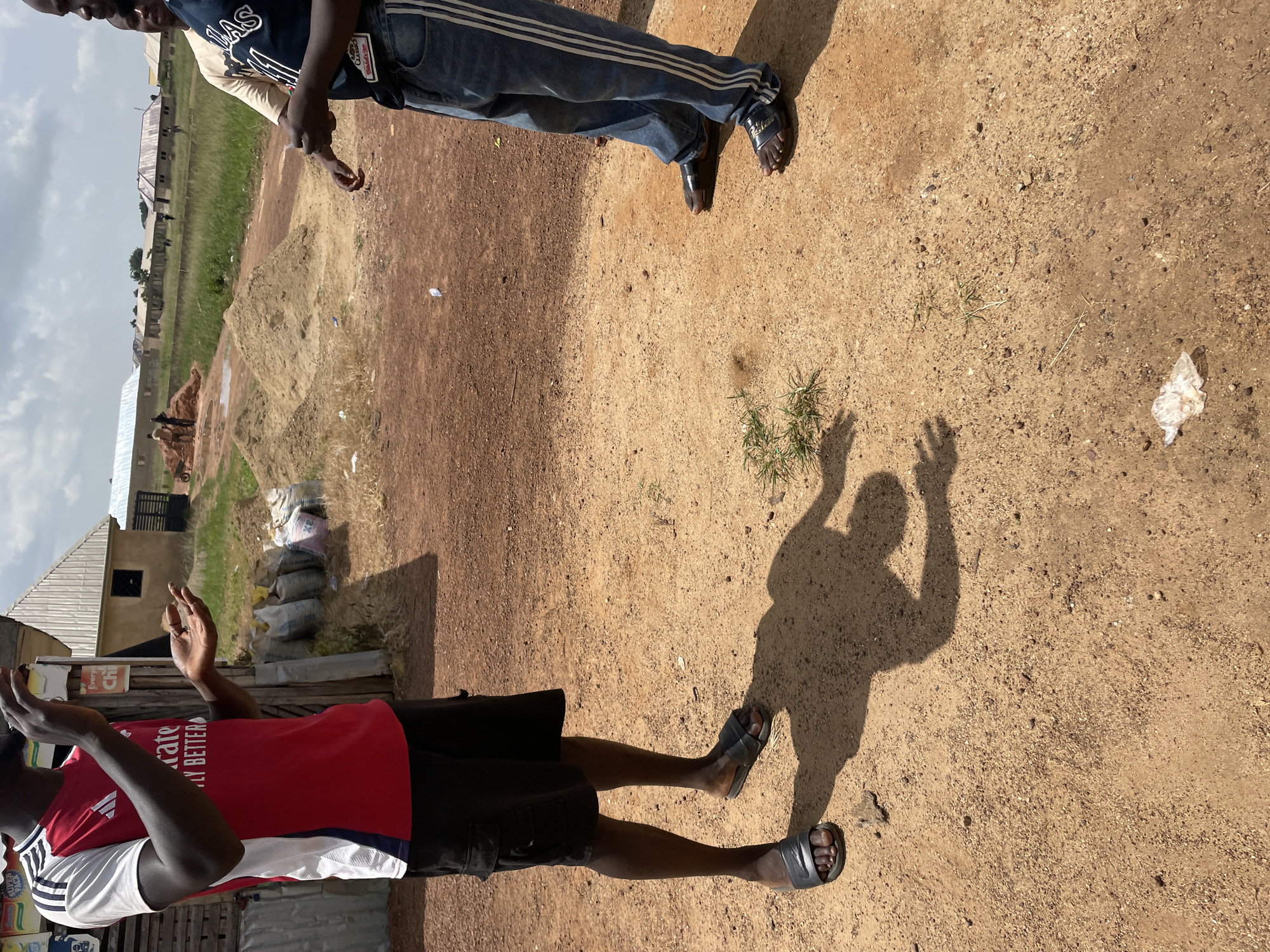 The image depicts an outdoor scene with two individuals, likely men, in Durumi, Nigeria. The foreground features one person wearing a red and white t-shirt with black shorts and black sandals. Their hands are raised in front of them. To the right, the shadow of a person with raised arms is cast on the dry, sandy ground. In the background, another person is partially visible, wearing a blue t-shirt and denim jeans with white stripes, also wearing sandals. The setting appears to be a dusty, open area, possibly a construction site or a similar unpaved locale, with piles of sand and some structures in the distance. Bags are stacked near a building, suggesting some form of commerce or storage. The sky is overcast, indicating a cloudy day. The overall impression is one of an everyday scene in a Nigerian community. No specific text is clearly legible.