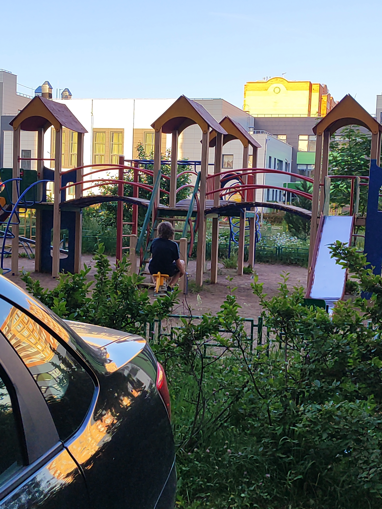 This image depicts a residential scene in Shchyolkovo, Russia, under clear skies with strong, warm sunlight, indicative of late afternoon or early morning. The foreground is occupied by the rear-left portion of a dark-colored vehicle, possibly black, with its surface reflecting the golden light of the sun and distorted images of surrounding buildings. Dense green foliage, including bushes and small trees, partially obstructs the view in the immediate foreground and along a green metal fence separating it from a playground.
The midground features a multi-component playground structure constructed primarily from wood, with reddish-brown pitched roofs on two tower-like sections. A wooden bridge connects parts of the structure, which also incorporates blue and red metal railings, ladders, and a white slide on its right side. On the playground's sandy ground, a single individual, seen from behind, is seated on a yellow, animal-shaped spring rider. The person wears a dark t-shirt and shorts, with dark, disheveled hair. Their presence is the only human element, and no interactions are depicted.
In the background, multiple multi-story buildings are visible. To the left and center, modern residential blocks with light-colored facades (white and grey) feature numerous windows. Dominating the right side of the background is a distinct yellow and orange building, brightly illuminated by the sun, showcasing a unique facade with a circular central feature and a distinctive roofline. Other buildings are present further back, some with green accents and visible roof details like vents and antennae against the pale blue sky. The overall environment appears to be a well-maintained urban residential area.