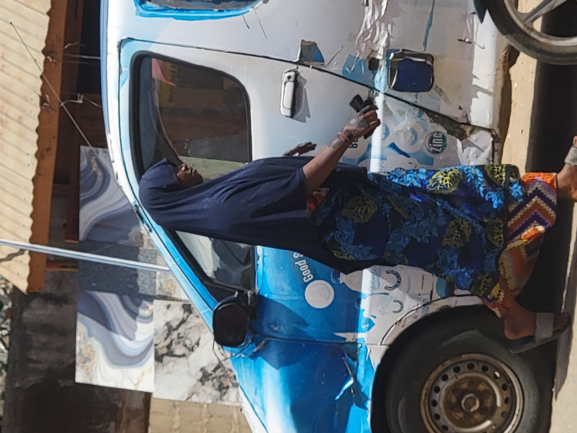 In Maiburiji, Nigeria, an adult female is sitting on a blue and white three-wheeled vehicle. She is wearing a dark blue headscarf and a blue floral dress. She is holding a dark object in her right hand. The vehicle has damage to its body. There are buildings in the background, and tiles are visible nearby.