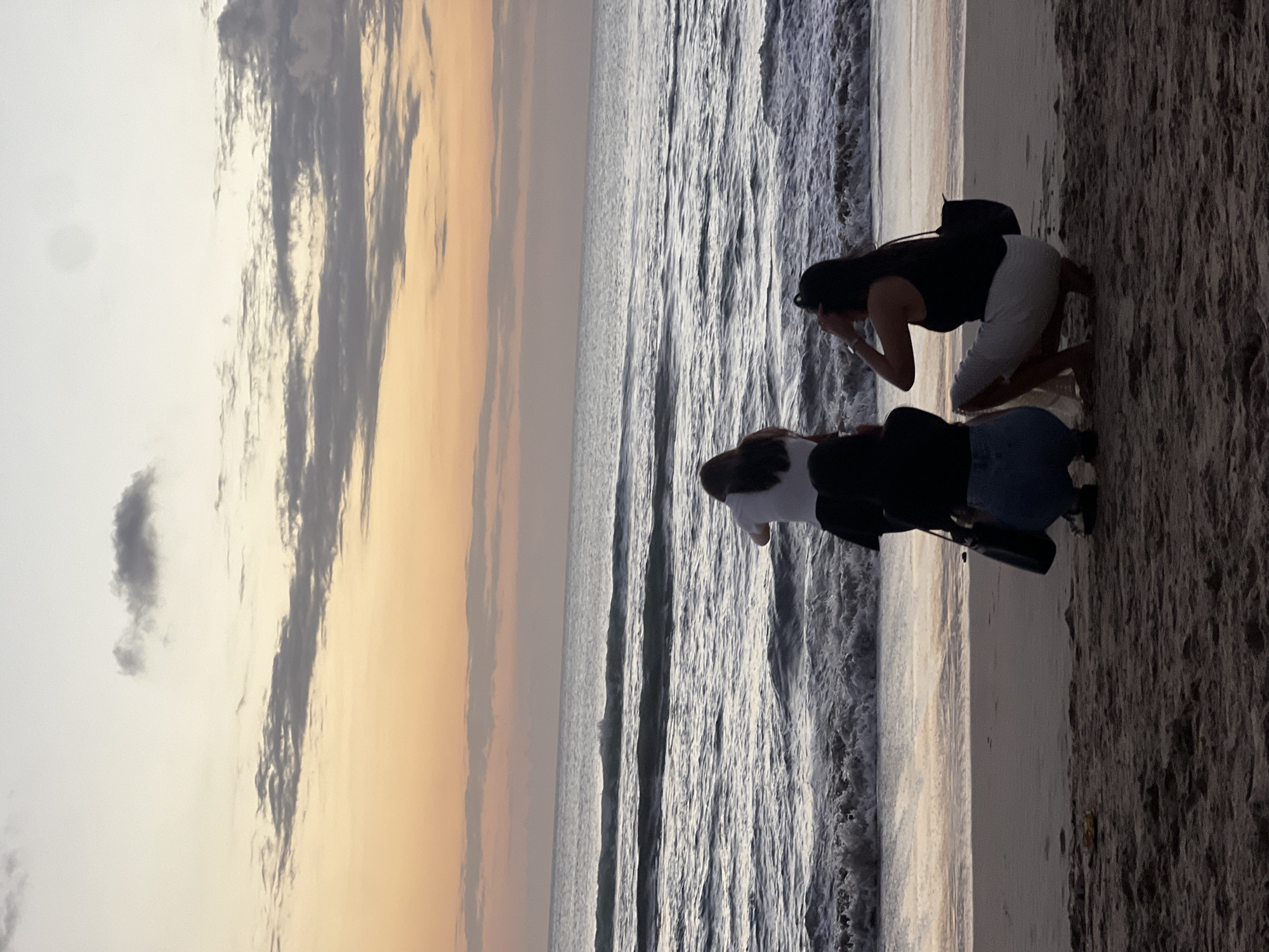 Three individuals are captured from behind on a sandy beach, facing the ocean during a sunset. The sky displays gradients of orange and gray clouds. Two individuals are crouching near the shoreline, while a third stands slightly further back. The waves are breaking on the shore. The scene suggests Kuta Beach, Indonesia, known for its sunsets.