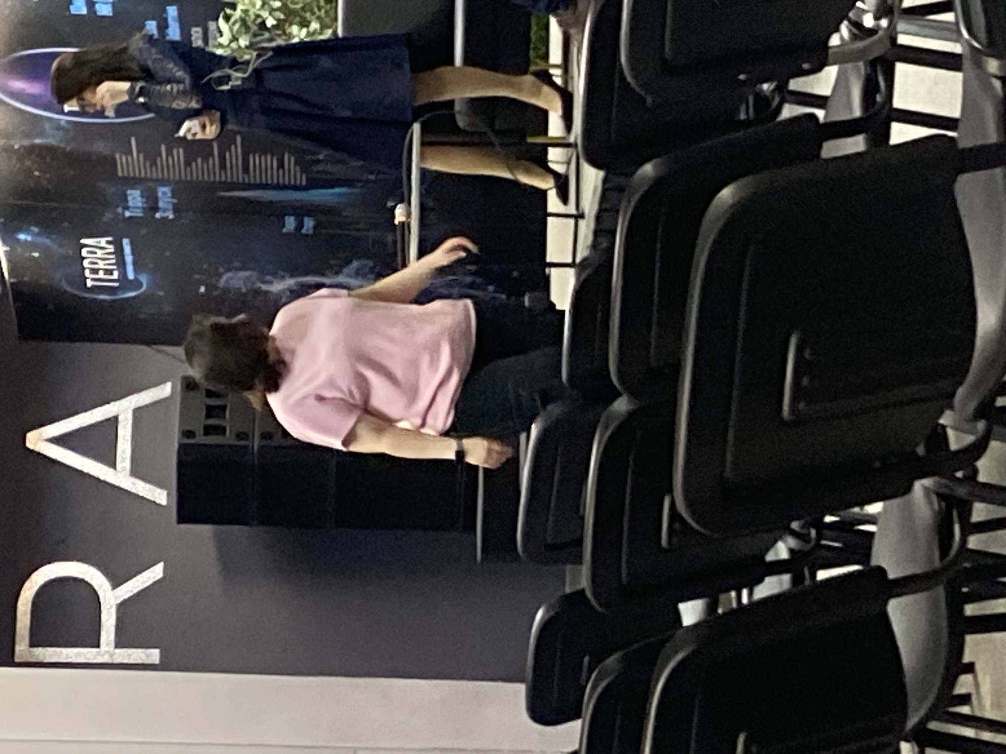 A woman in a pink t-shirt and dark pants stands facing left, her body angled away from the camera, on a stage. To her right, another woman in a dark blue dress and high heels stands facing the first woman, holding a phone in her right hand and gesturing with her left. Behind them, a backdrop features the word "TERRA" and other text. The foreground shows rows of empty black chairs. The scene appears to be an indoor event space.