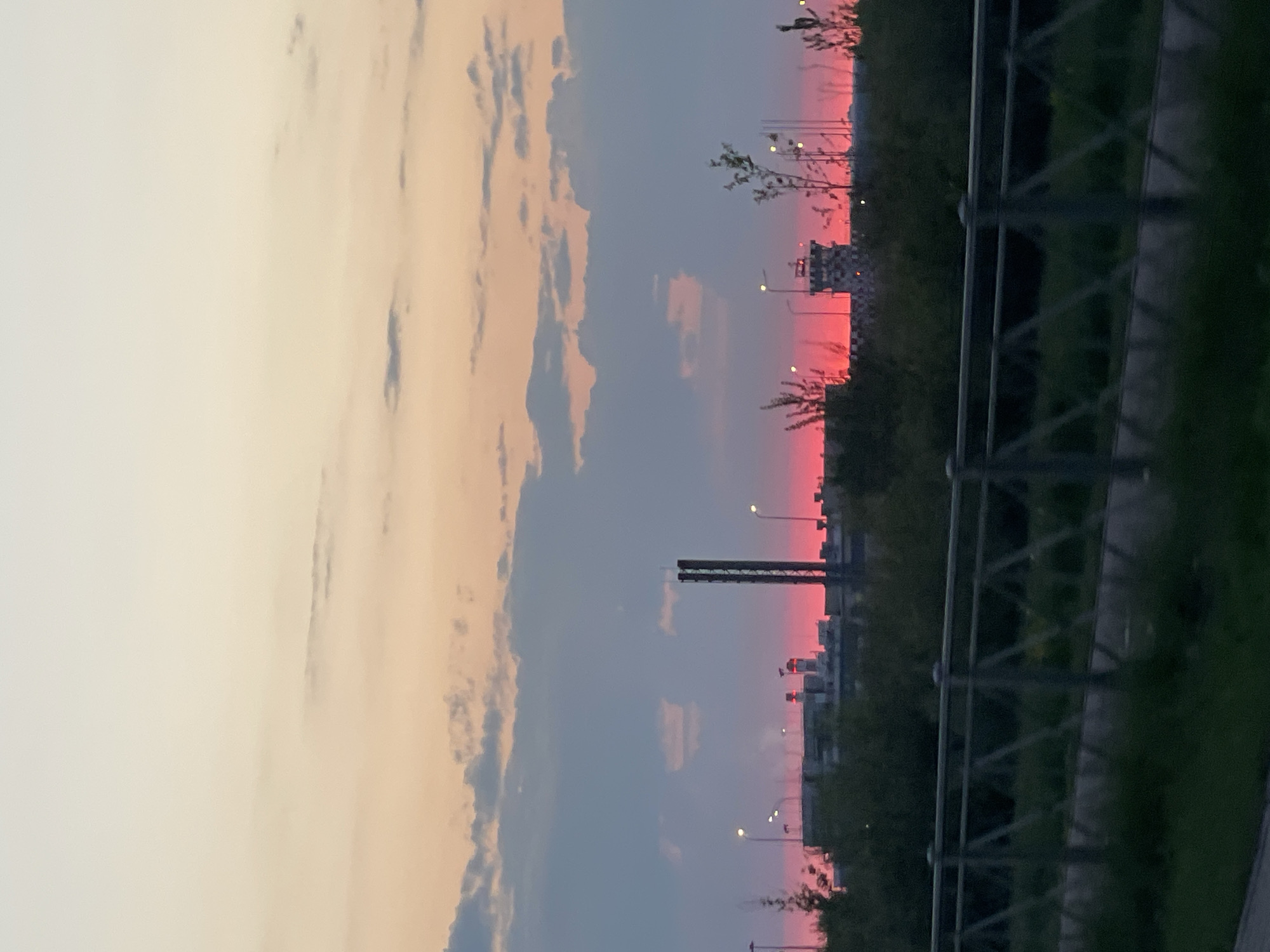 The image captures a vibrant sunset in Saint Petersburg, Russia. The sky displays a gradient of colors, transitioning from soft pink and orange hues near the horizon to a deep blue overhead. Clouds are scattered across the sky, adding depth and texture to the scene. In the foreground, a metal fence runs along the edge of a green area, possibly a park or roadside. Streetlights and silhouettes of buildings and structures are visible in the distance, suggesting an urban environment. The composition is somewhat tilted, which creates a dynamic perspective.
