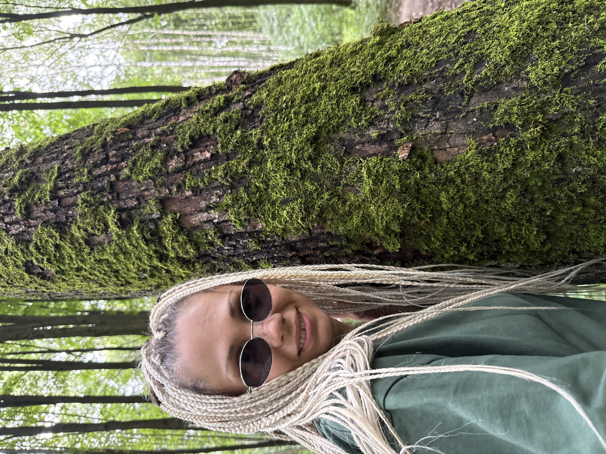 A woman with blonde braided hair and sunglasses smiles at the camera, standing next to a moss-covered tree in a forest. The trees are tall and slender, with green foliage. The woman is wearing a green hoodie. The location is the city of Dumino, Russia.