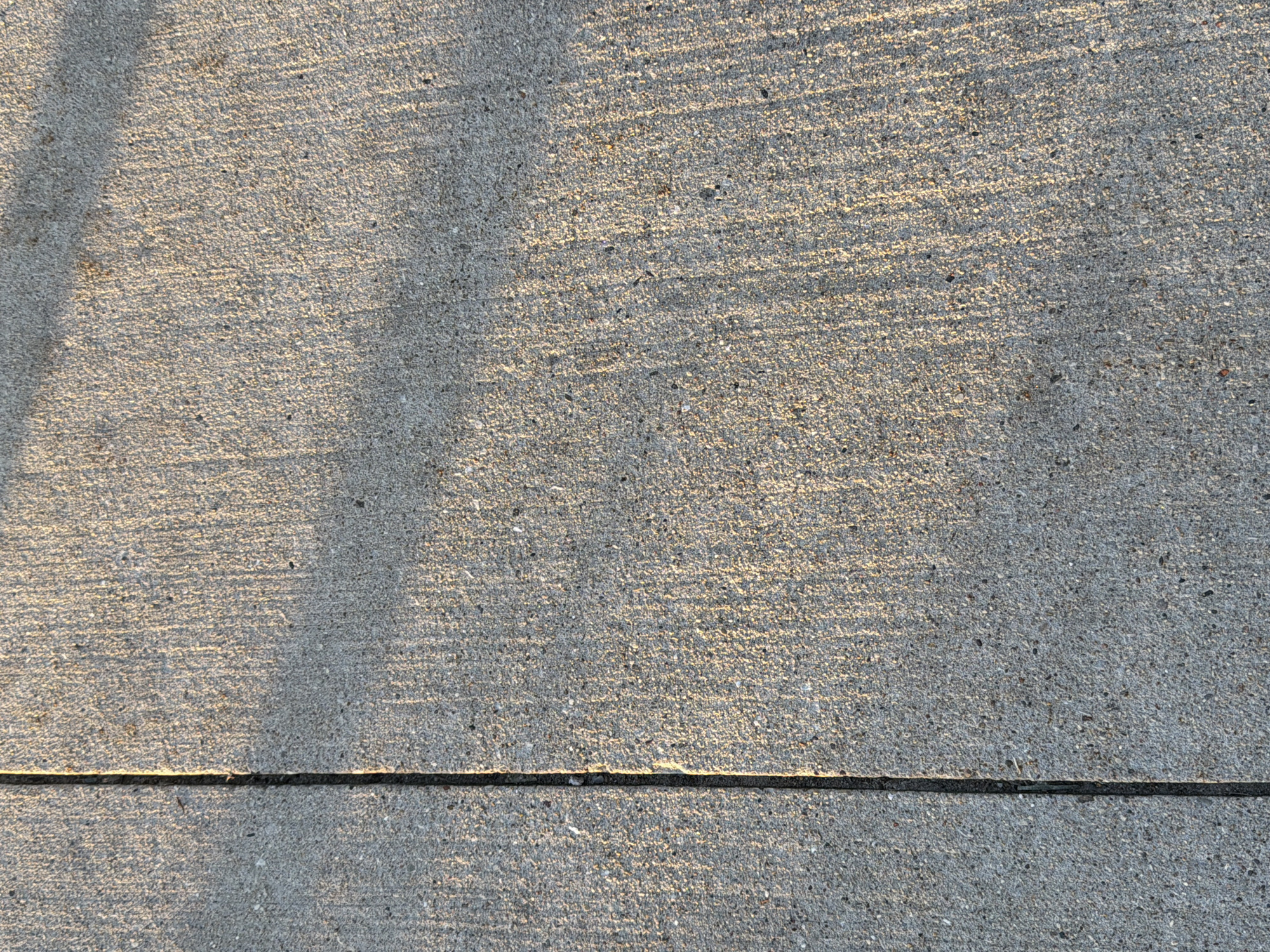 The image depicts a section of concrete sidewalk or pavement. The surface has a textured, slightly rough finish with a gray color. A dark, straight expansion joint runs horizontally near the bottom edge of the image. There are shadows cast across the surface, suggesting possible tree limbs or other overhead structures. The location is Morton Grove, United States of America. There are no people or other obvious objects in the scene.