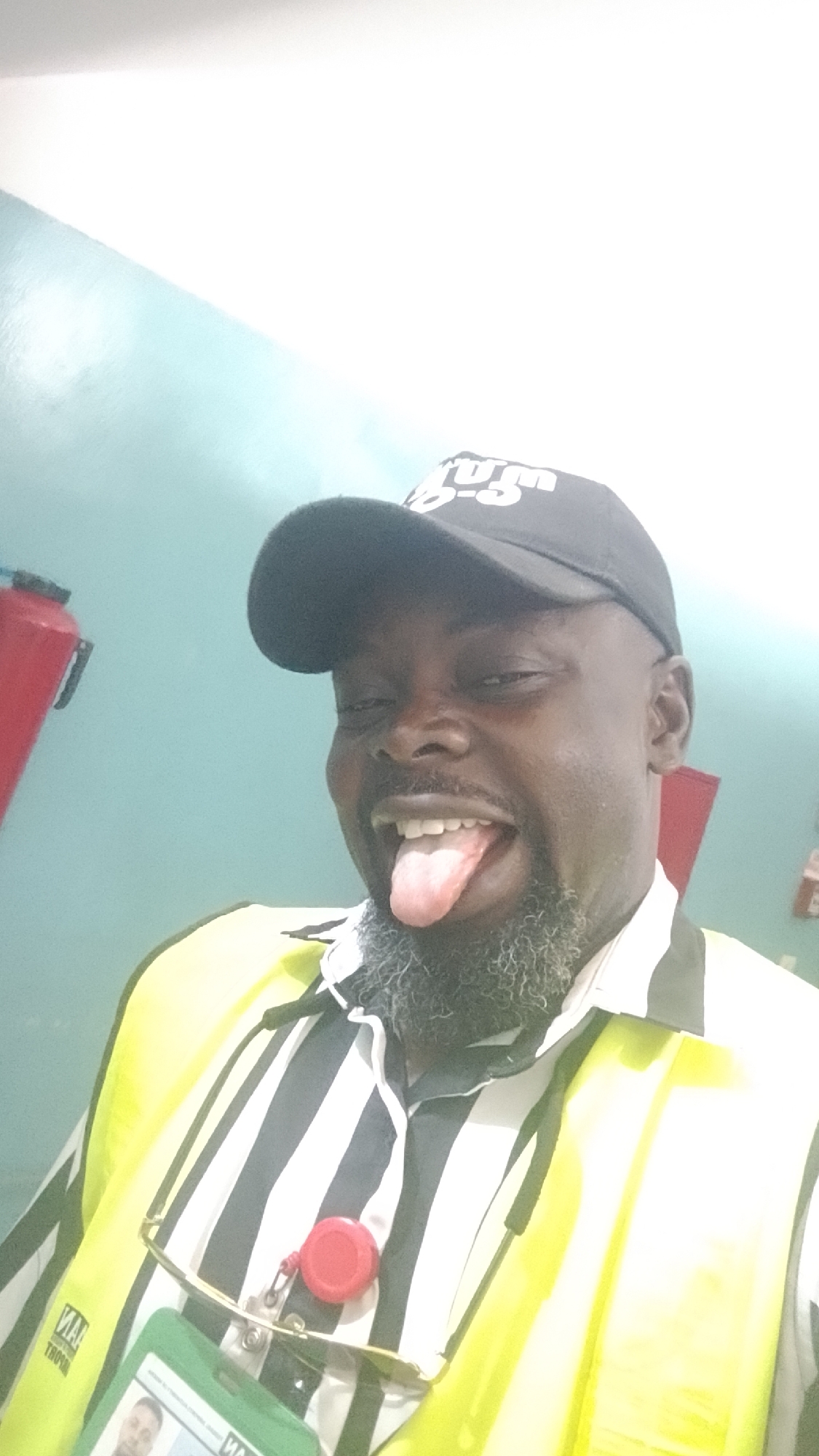 A man is posing for a selfie, sticking his tongue out. He is wearing a black baseball cap with white lettering, a striped black and white shirt, and a bright yellow safety vest. He has a dark complexion and a graying beard. An identification badge and eyeglasses are attached to his shirt. A red cylindrical object is also attached to his shirt. A red fire extinguisher is slightly visible on the left side of the image. The background consists of a light blue wall, and the overall lighting is bright. It is not possible to verify the location from the image.