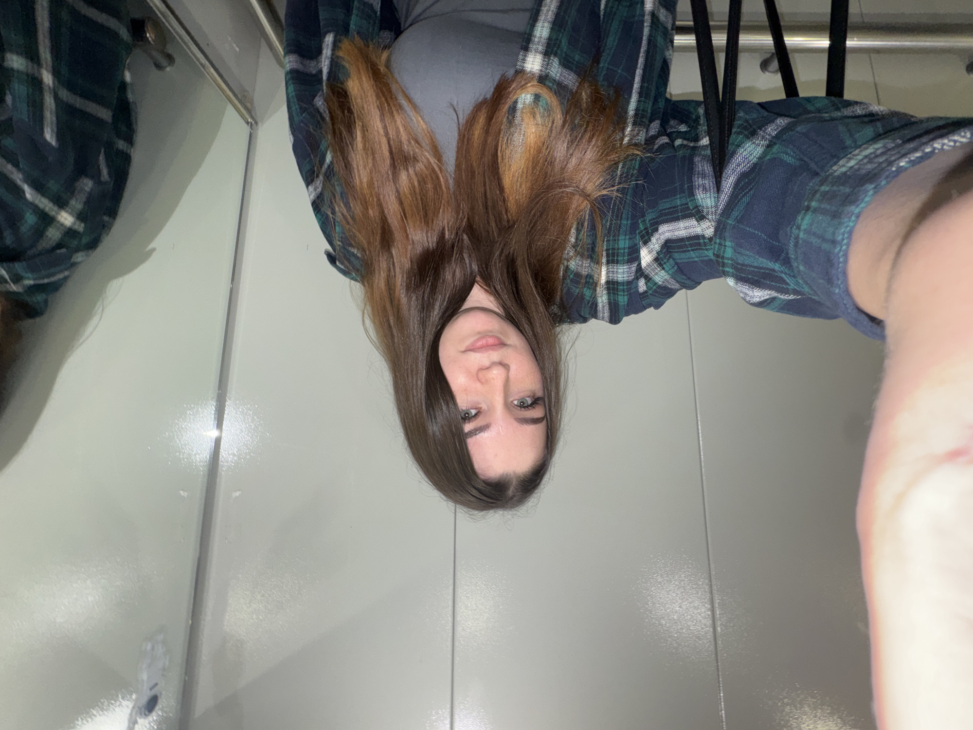 A young woman with long, brown hair is taking a selfie in what appears to be an elevator. She is wearing a dark blue and green plaid shirt over a grey top. The elevator walls are a light, reflective grey, and there are vertical seams dividing them. A metal handrail is visible along the bottom of the frame. In the reflection on the right side of the elevator, another person wearing a similar plaid shirt is partially visible. The overall environment is clean and appears to be a modern elevator. There is no specific visual information in the image that would allow for identification of the city as Perm, Russia.