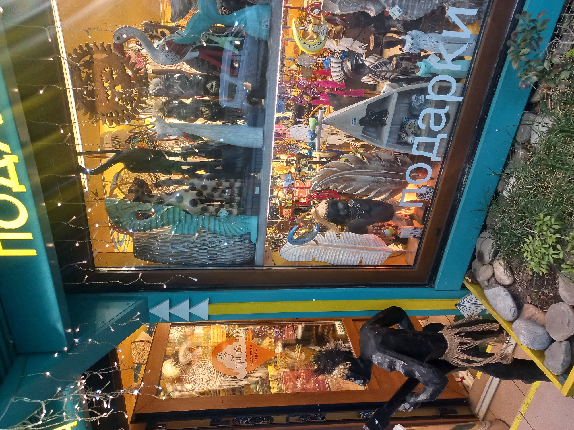 The image shows a storefront in Sochi, Russia. The store, called "Tri Neba" (Three Skies), sells exotic gifts. The window display includes a variety of handcrafted items, such as wooden figures of animals and celestial objects. Outside the store, there is a black wooden sculpture of a human figure. The store's facade is painted in blue and yellow. Decorations include strings of fairy lights.