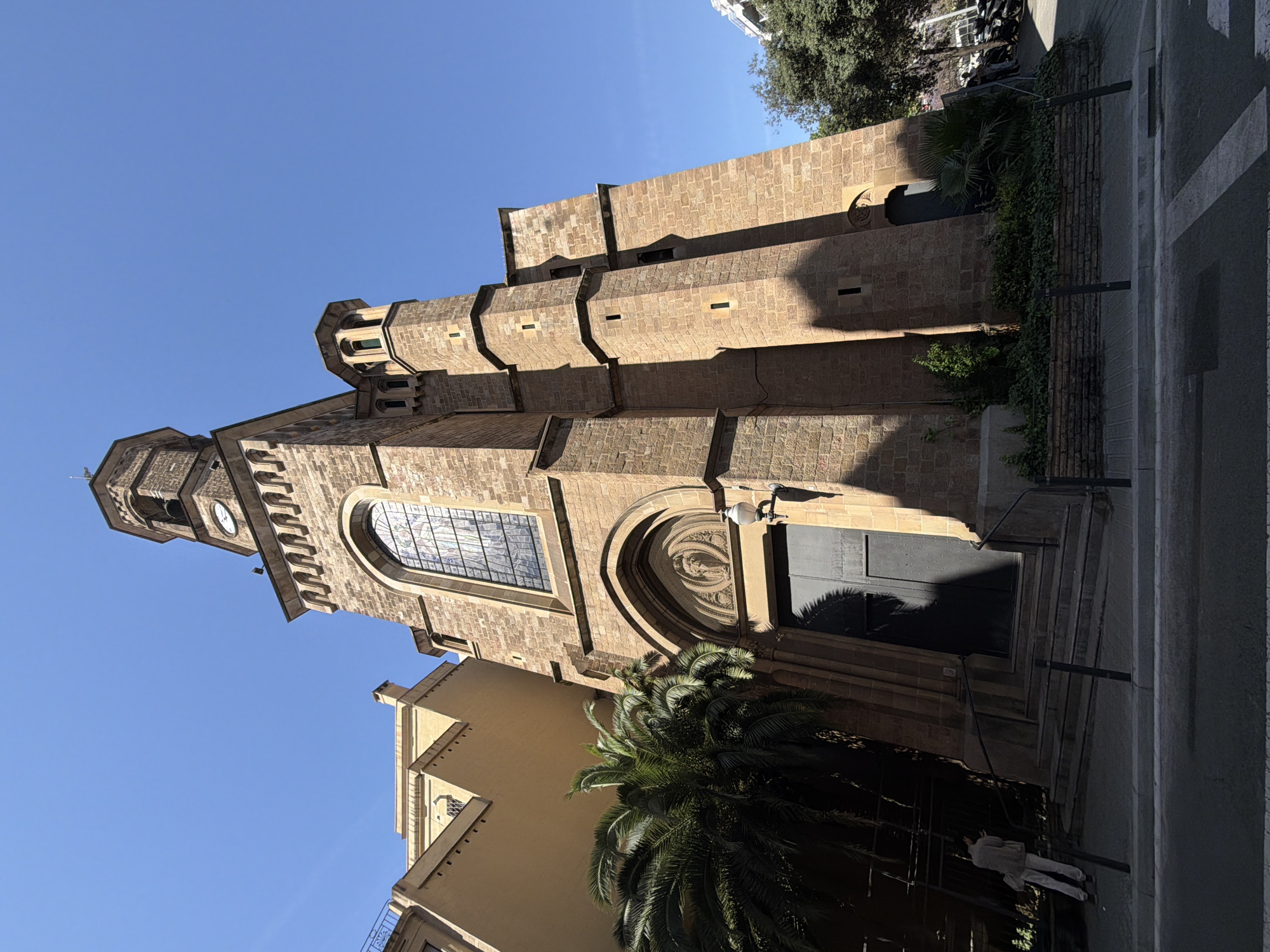 The image shows a low-angle view of a sandstone church, likely located in Barcelona, Spain. The church has a tall bell tower with a clock face, adjacent to the main building featuring a large arched stained-glass window and an ornate arched entrance with a closed, dark-colored door. The church's architectural style appears to be Gothic Revival. Palm trees grow near the building, adding a Mediterranean touch. The sky is clear and blue. A section of a building with light yellow walls is visible next to the church. Sidewalk is visible near the church entrance, with a pedestrian walking.