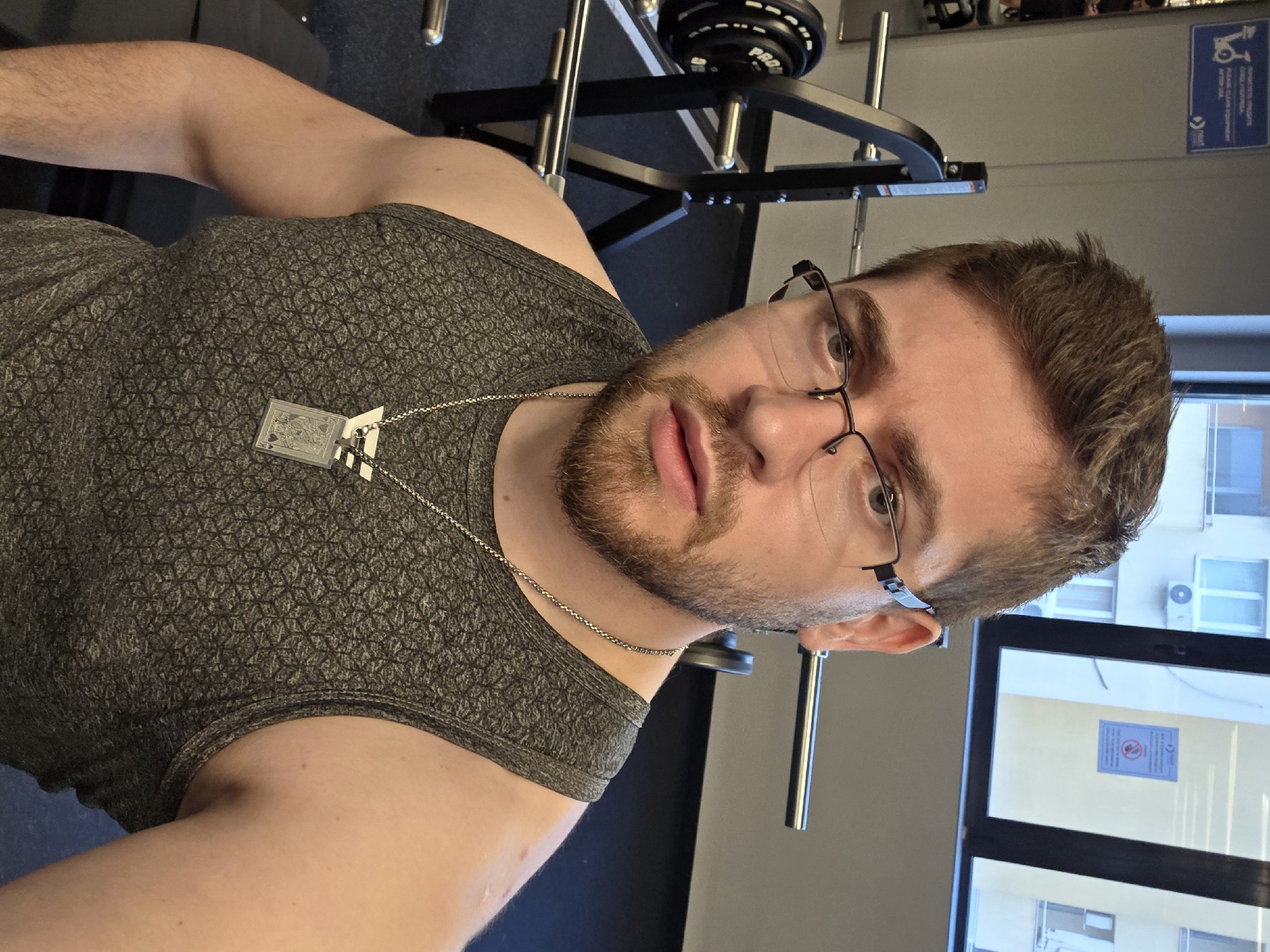 A close-up, slightly angled, selfie photograph shows a Caucasian man with short brown hair, a light beard, and glasses wearing a gray patterned tank top and a silver chain with a pendant. The man is looking directly at the camera with a neutral expression. Behind the man is weightlifting equipment, including a rack and weights. Through a window, a building exterior is visible, suggesting the location is in an urban setting. Based on the window view, the scene potentially located in the city of Plovdiv, Bulgaria. A blue sign is visible on the wall behind the man and the weightlifting equipment. The floor is dark gray.