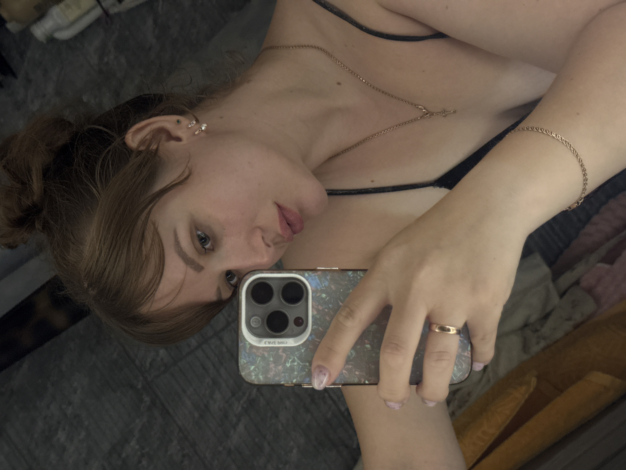 A young woman is taking a mirror selfie in what appears to be a bedroom. She has her hair up in a bun and is wearing a dark-colored spaghetti strap top. She is also wearing jewelry including a necklace with a cross pendant, earrings, a ring on her left hand, and a bracelet on her right wrist. She is holding a phone in her left hand, which is reflecting in the mirror. The background is blurred, but it appears to be a wall with some objects on a shelf. The woman is looking at her reflection in the mirror. There is no indication of the location being in Chaikovsky, Russia.