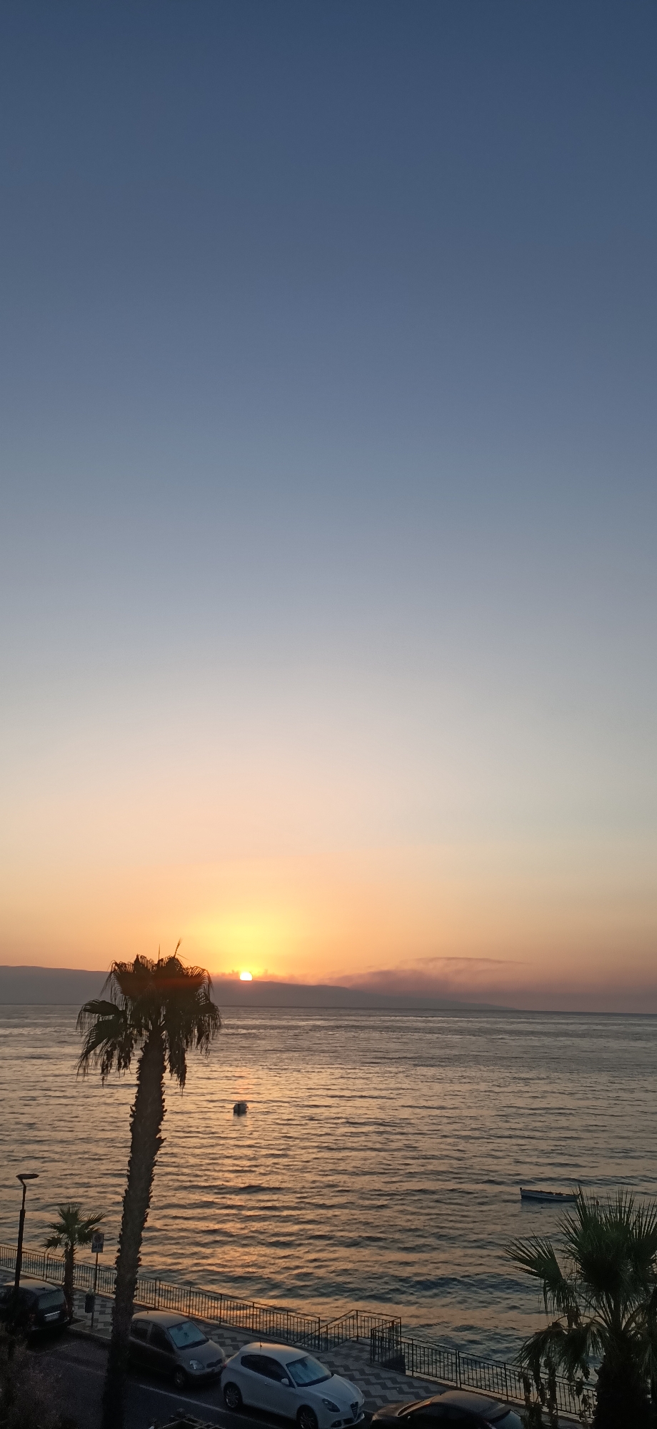 The image depicts a sunset over a body of water, likely a sea, with a coastline and urban elements in the foreground. The sky transitions from a clear, deep blue at the top to a soft gradient of pale blue, peach, and orange near the horizon where the sun is setting. The sun itself is partially obscured by a thin layer of clouds and appears as a bright, glowing orb.
The water is calm with gentle ripples reflecting the warm hues of the sky. In the distance, a dark, mountainous landmass is visible along the horizon.
In the foreground, a tall palm tree stands prominently on the left side of the image, its fronds silhouetted against the sky. Below the palm tree, a paved walkway or promenade runs parallel to the water, bordered by a metal railing. A street is visible behind the walkway, with several parked cars, including a white hatchback, a dark sedan, and another dark vehicle. Streetlights and smaller palm trees are also present along the street.
Further out on the water, there are two small boats. One is closer to the shore and appears to be a dark rowboat. Another object, possibly a buoya, is visible closer to the palm tree. The overall atmosphere of the image is serene and peaceful, characteristic of a sunset scene. The location appears to be a coastal urban area.