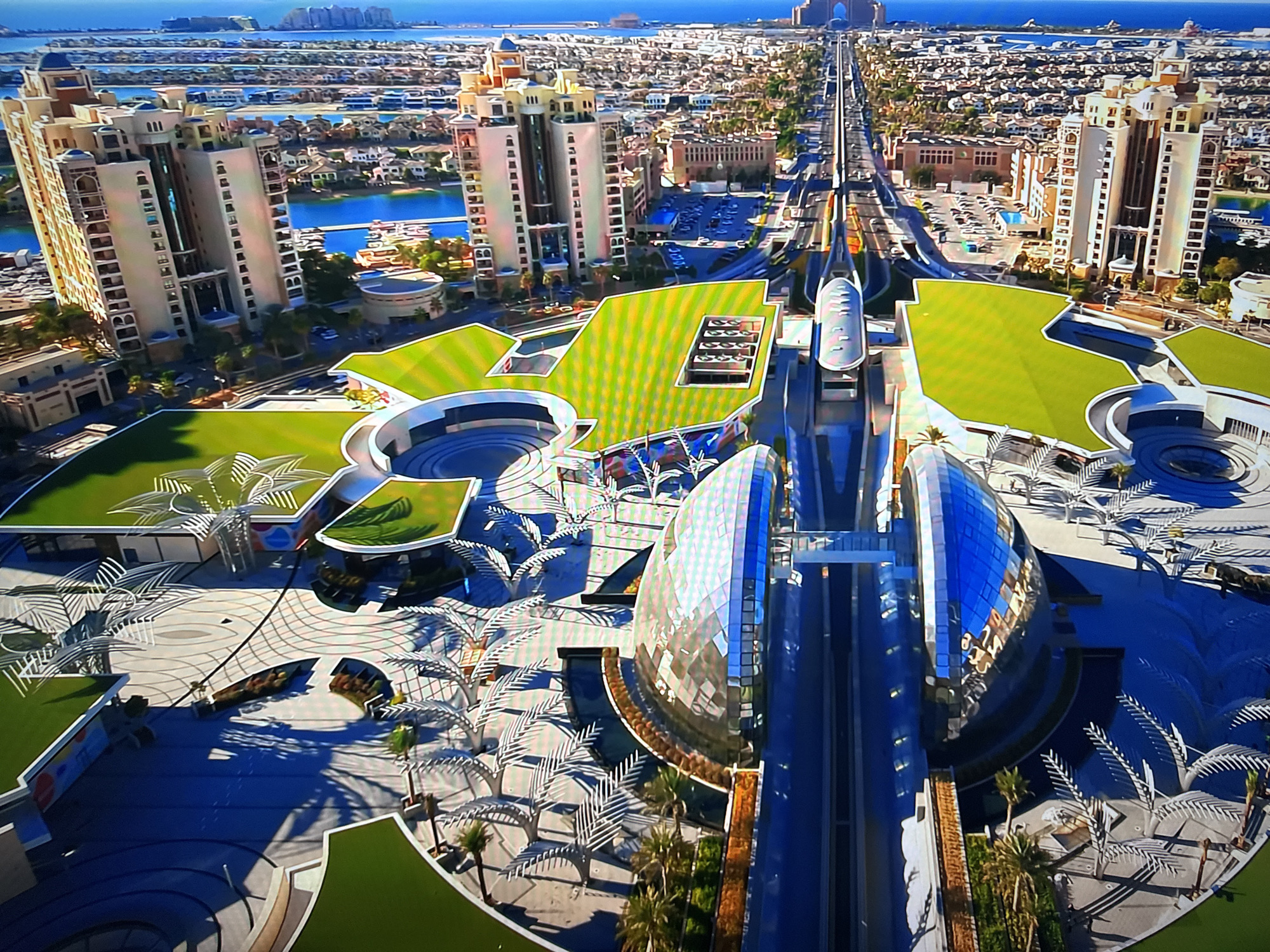 The image is an aerial view of a modern city with distinct architecture. In the foreground, there is a large, contemporary complex featuring curved, glass-domed structures and green, angled roofs. White, stylized palm trees and pathways are visible in the open spaces. A monorail track runs through the complex, with a train visible on it.
Behind the complex, a wide avenue stretches into the distance, lined with buildings on either side. These buildings are a mix of tall, apartment-style towers with light-colored facades and more traditional, lower-rise structures. Beyond the avenue, canals or waterways are visible, reflecting the blue sky. In the far distance, a sprawling urban landscape with numerous buildings and a prominent man-made island structure can be seen, with the ocean beyond.
The location is not in Burgas, Bulgaria. This appears to be a development in Dubai, likely on Palm Jumeirah, given the distinctive architecture and the presence of the monorail system.