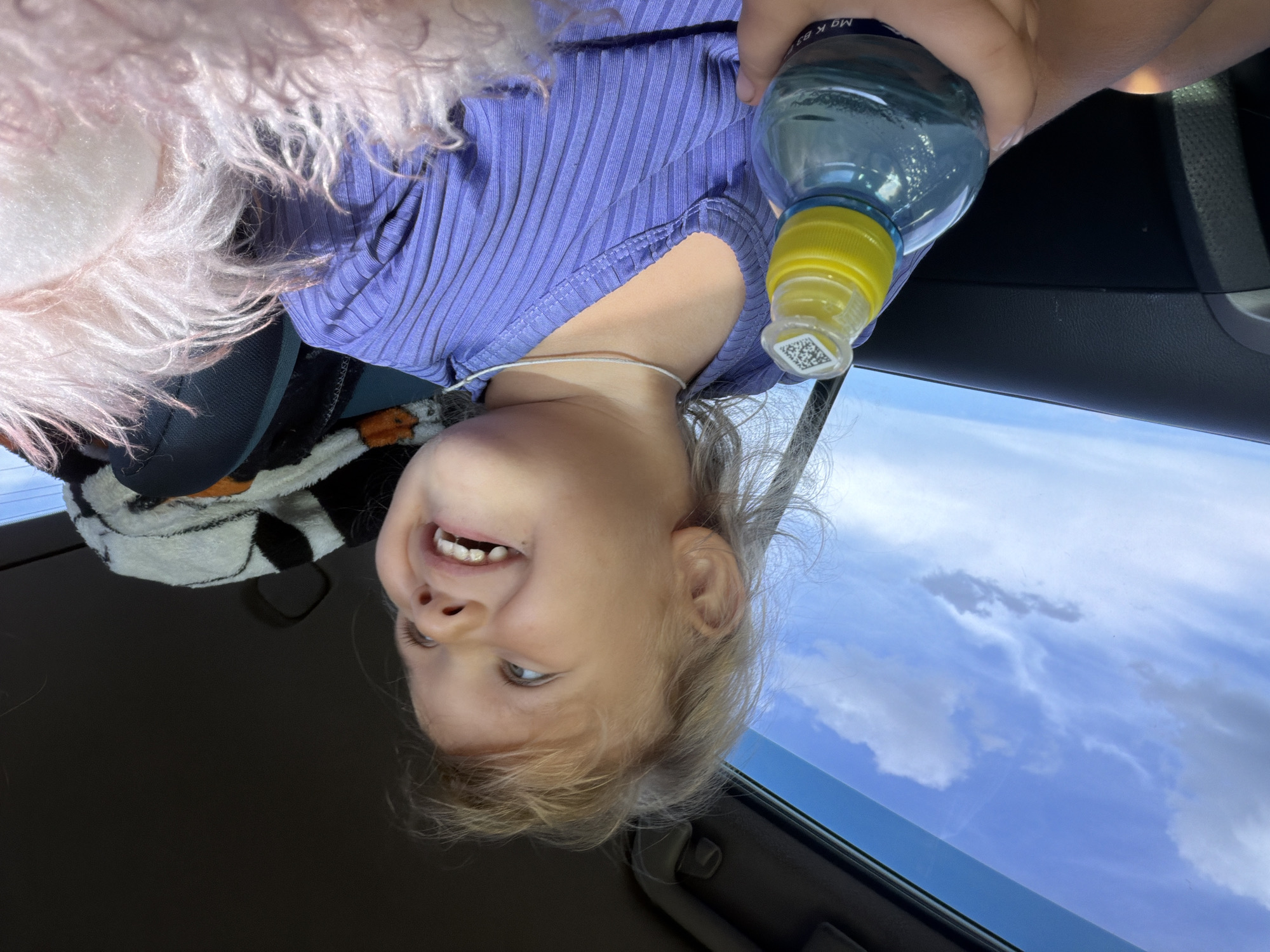 A young child with blonde hair is in a car, holding a blue water bottle with a yellow cap. The child is wearing a purple ribbed shirt and looking out the window with a wide smile. The sky is visible through the car window, with scattered clouds. The child's right hand is visible on the water bottle. In the background, a black and white striped blanket and a pink fuzzy object are visible. The overall impression is one of a happy child on a journey. The image is captured from a slightly elevated perspective.