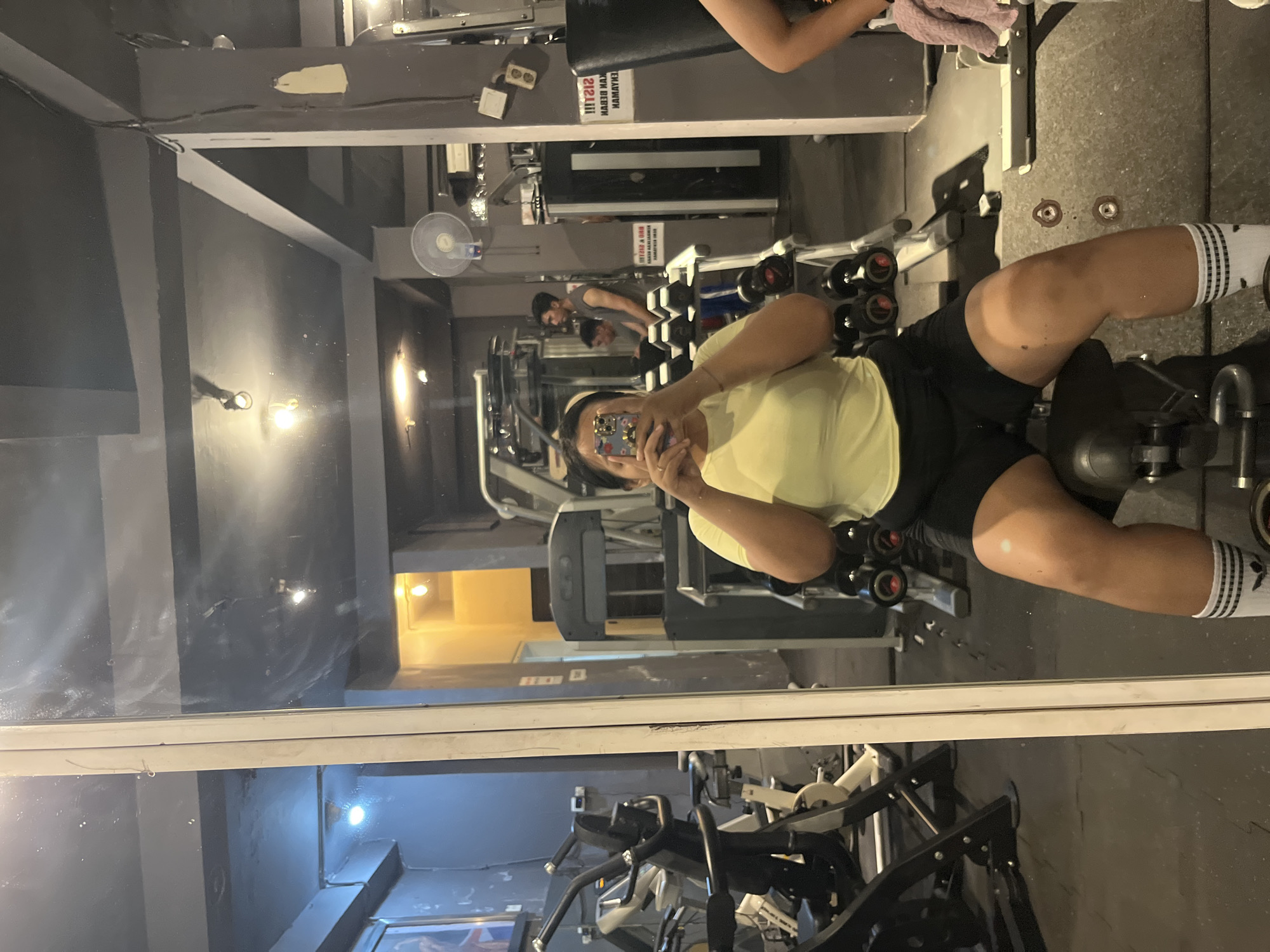 A person is taking a selfie in a gym mirror. The person is wearing a yellow t-shirt and black shorts, with white socks. They are sitting on a weight machine. In the background, other people are visible, as well as gym equipment such as treadmills and weight racks. The gym appears to be located in Denpasar, Indonesia, based on a sign in the background with Indonesian writing.