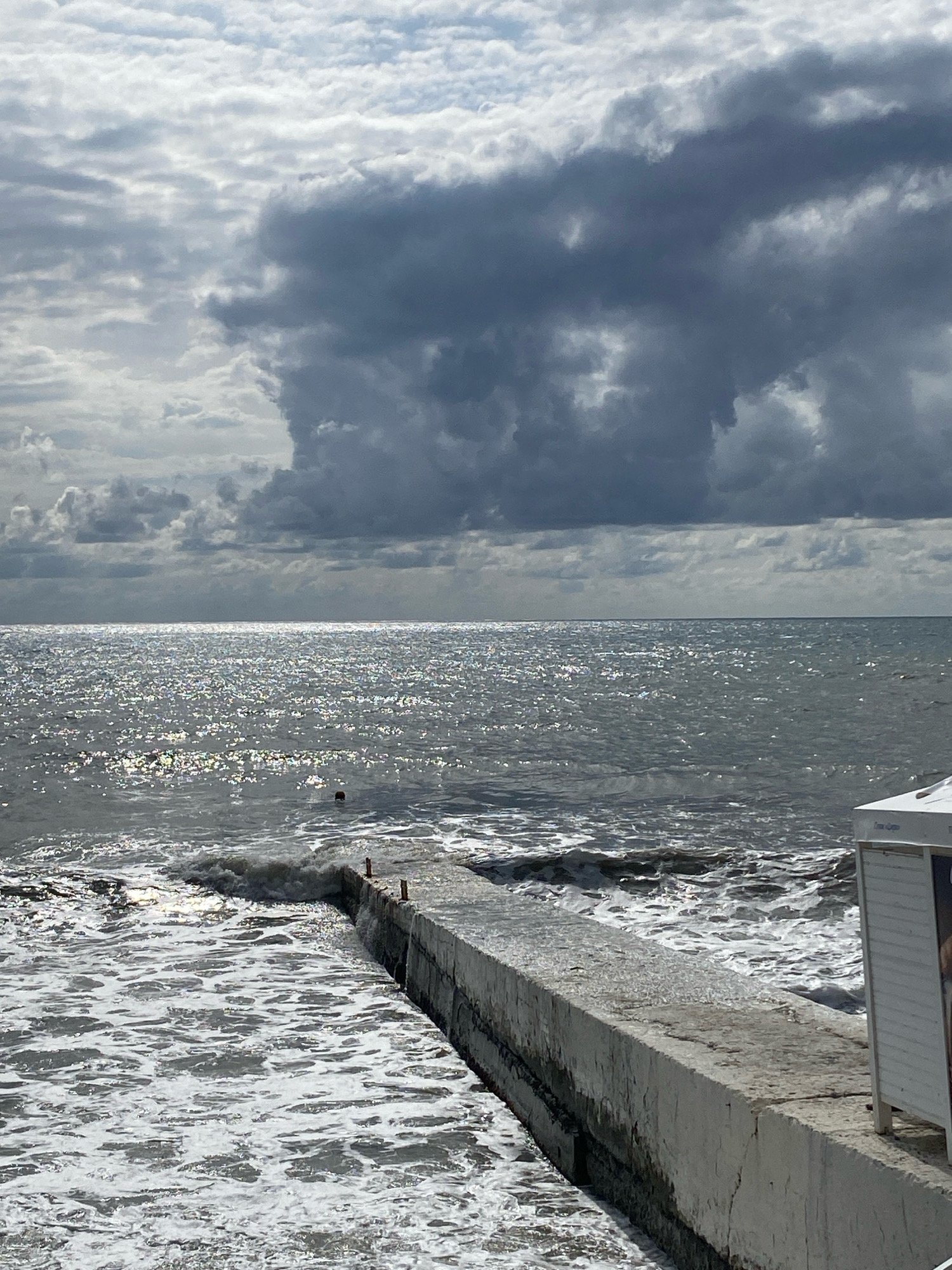 The image depicts a cloudy day at the sea in Sochi, Russia. A concrete pier extends into the choppy, sun-dappled water. Waves crash against the pier, creating white foam. In the background, the sky is filled with dramatic dark clouds, contrasting with patches of lighter clouds and the bright reflection of the sun on the water. To the right of the pier, a small white structure, possibly a changing room or kiosk, is partially visible. No people are clearly visible in the scene.