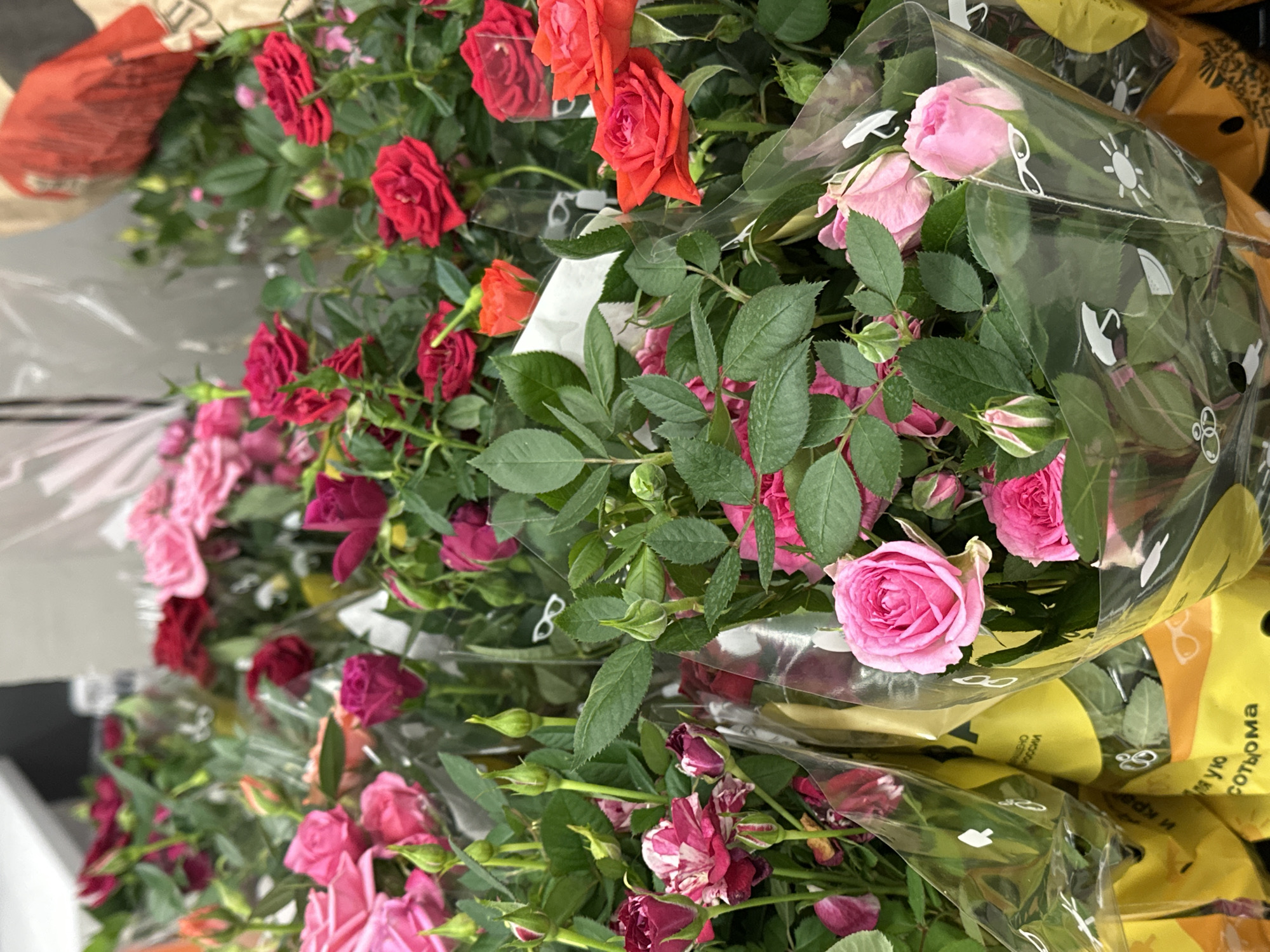 The image shows a display of bouquets of miniature roses, likely in a store located in the Бараўлянскі сельскі Савет area of Belarus. The bouquets feature a variety of colors, including red, pink, and burgundy. They are wrapped in clear plastic with yellow packaging visible beneath, some of which has white graphics and text in Cyrillic. The roses are arranged closely together, suggesting they are being sold. The display is situated indoors, potentially in a supermarket or flower shop.