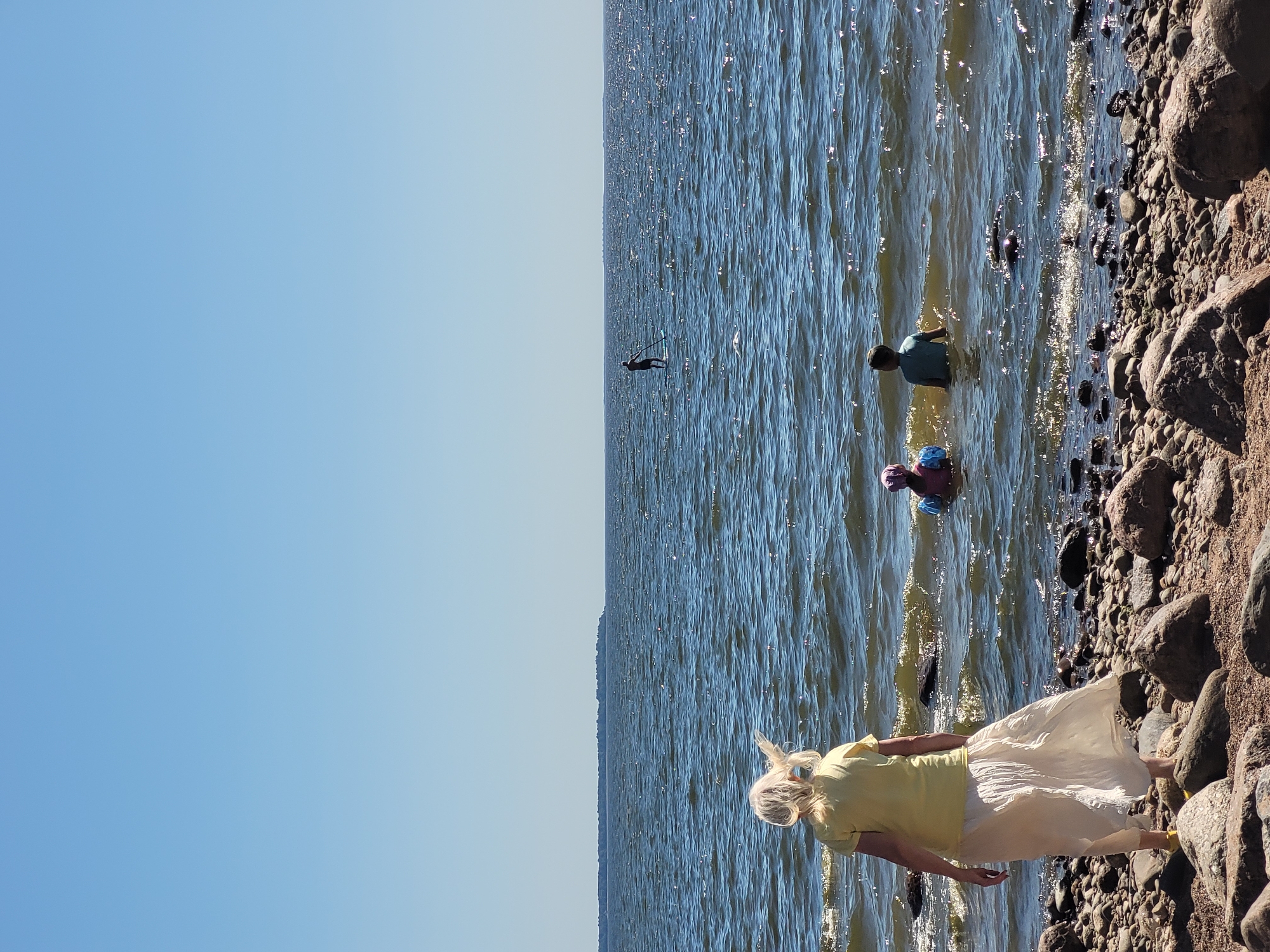 In Repino, Russia, on a sunny day, a woman with blonde hair, wearing a yellow t-shirt and a long, flowing white skirt, stands on a rocky shore facing a calm, sun-dappled body of water. Two children, one wearing a pink hat and the other a blue shirt, are wading in the shallow water. In the distance, a lone figure stands on a paddleboard in the expansive water. The horizon is marked by a thin strip of land under a clear blue sky.