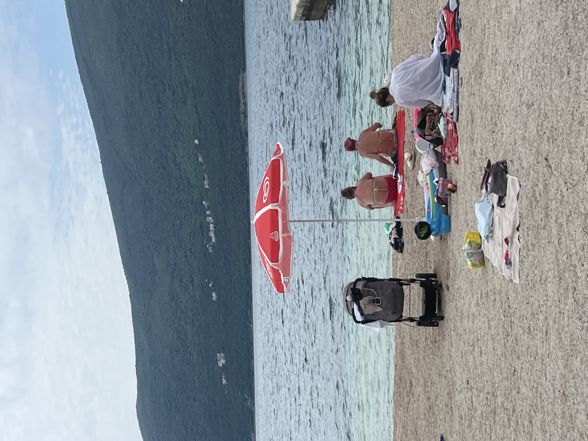 This daytime image captures a tranquil beach scene in Baošići, Montenegro, with a forested mountain, calm water, and several people relaxing. The view is predominantly split between a large, dark green, tree-covered mountain range on the left and the water and beach on the right. The sky above the mountains is light blue with scattered white clouds.
In the foreground, a light-colored, possibly pebbly, beach runs along the right side of the frame, meeting the placid, light blue-green water. Closer to the water's edge, a red beach umbrella, bearing the distinct "Magnum" logo (a stylized heart or 'M' mark), is open, providing shade. To the right of the umbrella, a dark gray and black baby stroller is parked on the beach.
Several individuals are present near the water. One person, seen from the back with their hair in a bun, is wearing a white top and appears to be attending to belongings spread on a red mat. Two other individuals, also viewed from behind, are sitting or crouching in the shallow water, wearing red swimwear. Various personal items, including towels, clothing, and bags, are scattered on the beach around the people and the stroller. A blue inflatable object and some yellow packaging are also visible.
The scene conveys a sense of peaceful leisure, with people enjoying the coastal environment. The time of day appears to be late morning or early afternoon, with soft, diffused sunlight.