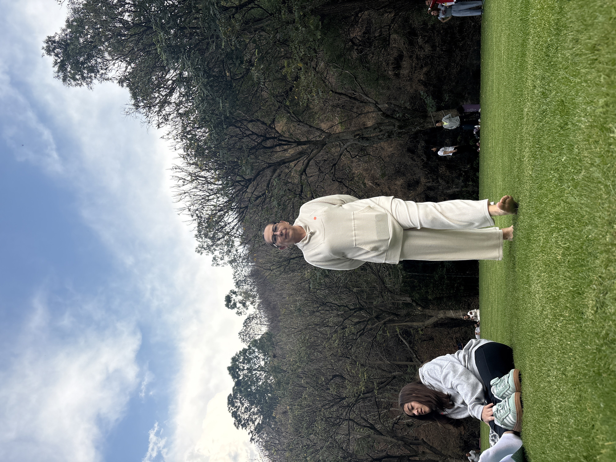 The image shows an outdoor scene, likely in Santa Ana Acozautla, Mexico, featuring a group of people on a grassy field surrounded by trees. A man in white clothing stands with his arms behind his back. Another person, a woman, is kneeling and looking down. A group of people stand in the background. The sky is visible with clouds.