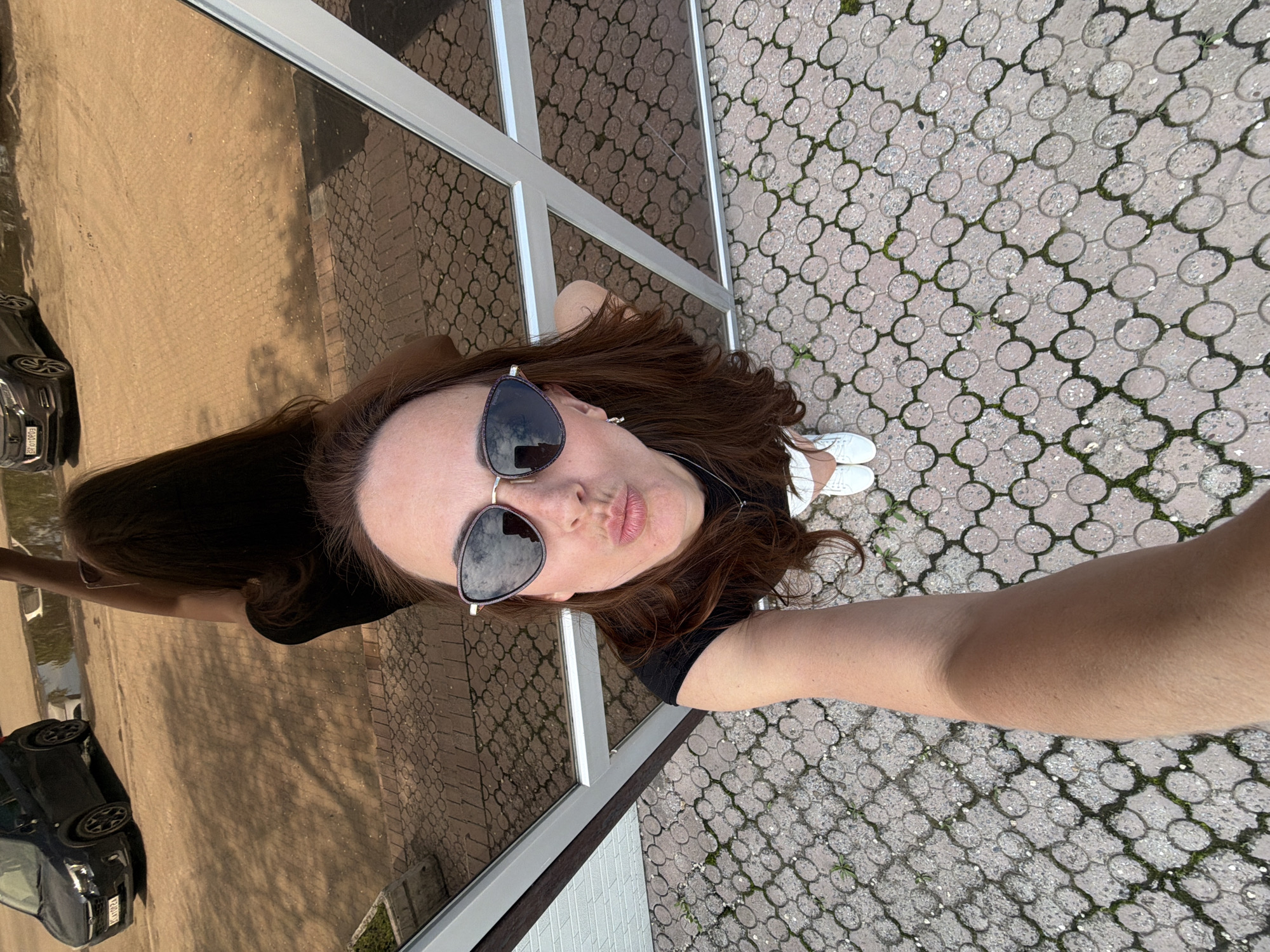 A woman with long brown hair and sunglasses is taking a selfie outdoors. She is wearing a black t-shirt and white sneakers. The background shows a reflection of a dark car and a paved area with a circular pattern. There are metal framing elements visible in the foreground. The environment appears to be a public space, possibly near a building with reflective surfaces.