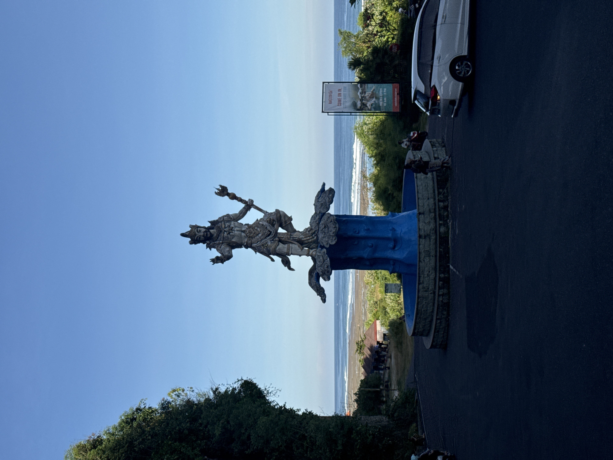 The image depicts a stone statue of a deity holding a trident, elevated on a blue pedestal with ornate stone carvings. The statue is positioned alongside a coastal road in Kuta Selatan, Indonesia. The Indian Ocean stretches to the horizon, meeting a clear blue sky. Vegetation grows along the coastal road and on a cliff face. A road runs parallel to the coast with a white vehicle parked on the side. A sign advertising "Water Blow" is visible. There are some people standing near the statue. The viewpoint is from ground level, looking upwards.