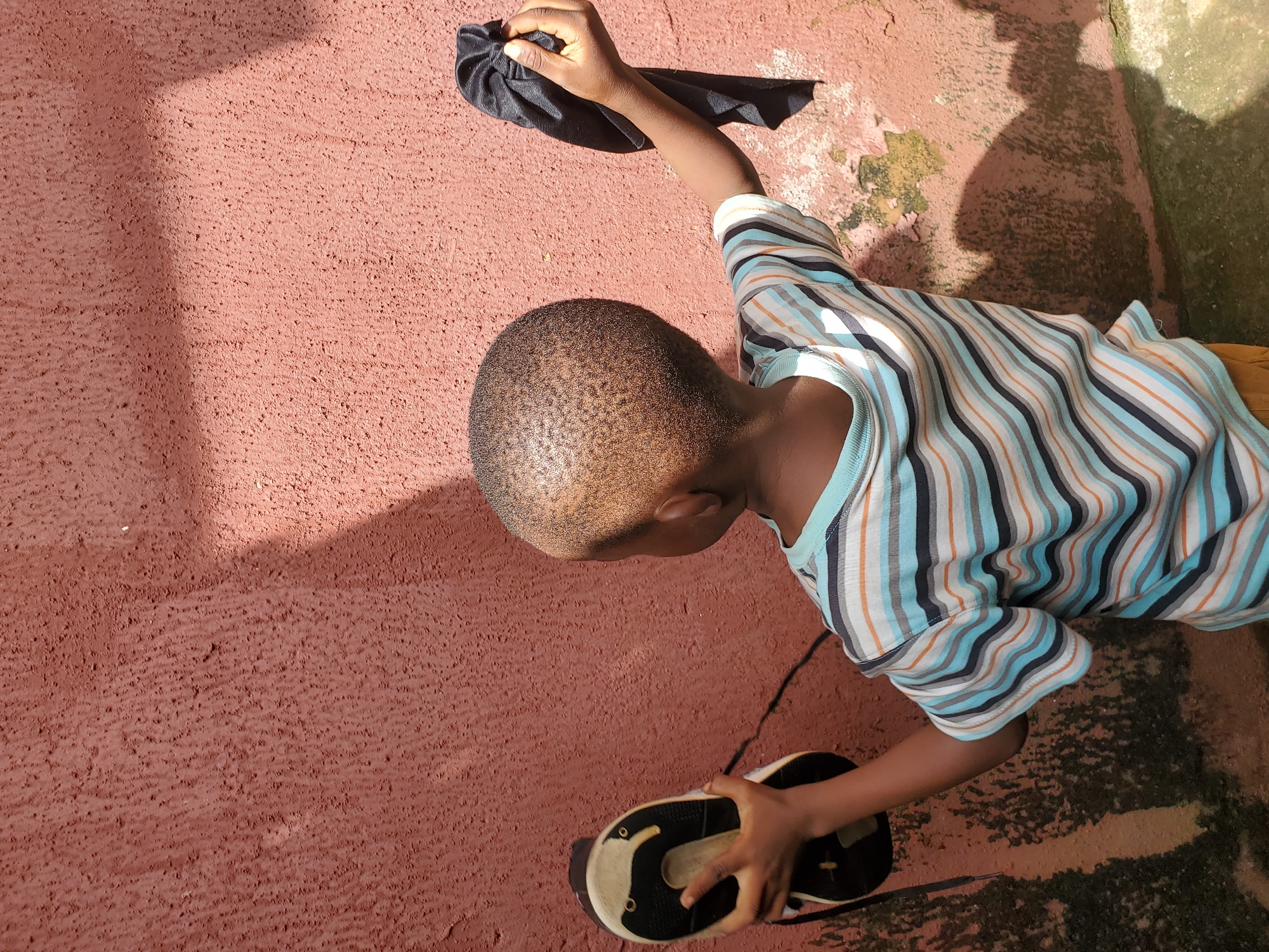 A young child, viewed from behind, stands facing a rough, salmon-pink wall in what appears to be an outdoor setting. The child, with a closely cropped haircut, wears a horizontally striped t-shirt in shades of blue, white, black, and orange. In their right hand, the child holds a black piece of cloth, bunched up and grasped at the top. In their left hand, the child holds a black and white shoe, its sole facing outwards. The wall exhibits signs of wear, with patches of discoloration and mossy growth, particularly in the lower left corner. Sunlight casts distinct shadows on the wall, indicating a bright, sunny day. The ground below the wall is visible in the lower portion of the frame, appearing to be a concrete surface with some debris. This scene is located in Jos, Nigeria.
