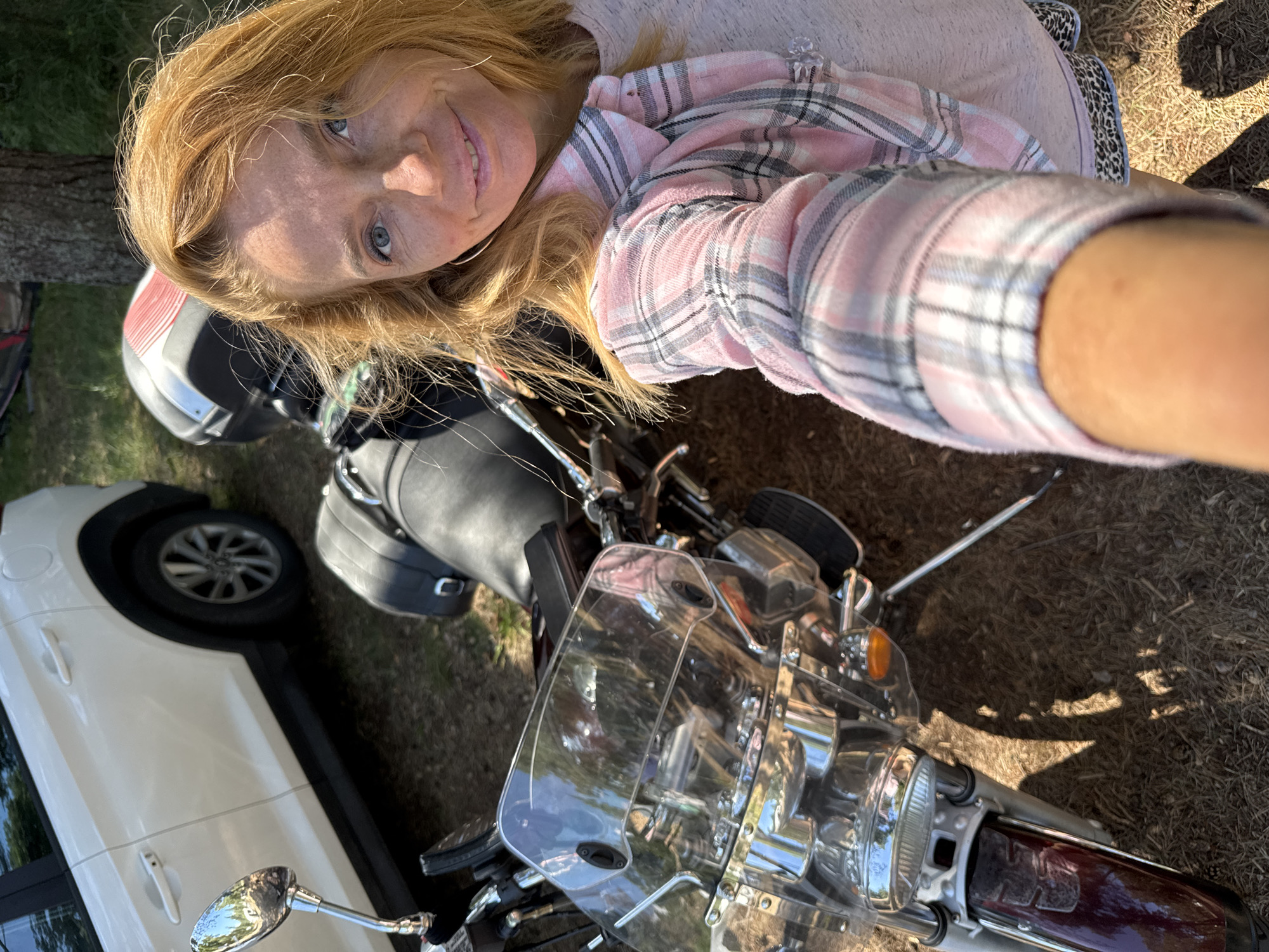 In Zelenaya Roshcha, Russia, a woman with auburn hair and blue eyes is captured in a selfie. She is wearing a pink and white plaid shirt over a light pink top and leopard print shorts. To her left, a motorcycle with a large clear windshield and a red and black storage compartment is parked. Behind the motorcycle, a white SUV is visible. The scene is set outdoors on a sunny day, with dry grass and shadows suggesting a natural or park-like environment. The woman is smiling directly at the camera, holding it out to take the photo.