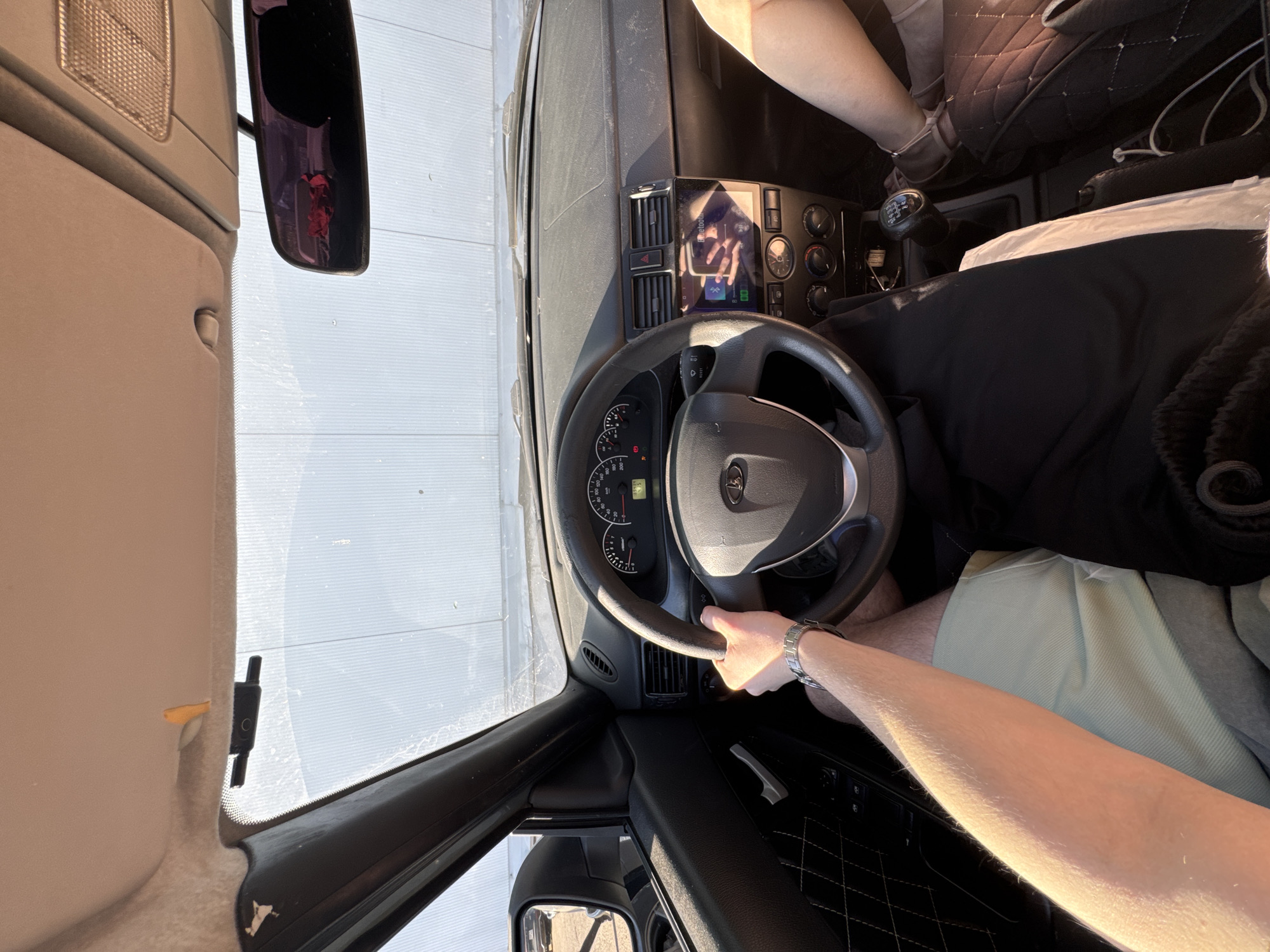 The image depicts the interior of a Lada car, from the driver's perspective. The driver is holding the steering wheel with their left hand, with a watch visible on the wrist. The dashboard is visible, featuring a touchscreen entertainment system. The gear stick is located to the right of the steering wheel. The passenger seat is covered in a black, quilted material. Outside the car, through the windows, a metallic facade is visible, suggesting the car is in a urban setting. Based on the presence of Lada car, the image is likely taken in Russia. Given that Perm is the only city mentioned, it is inferred that the photo was taken in Perm, Russia.