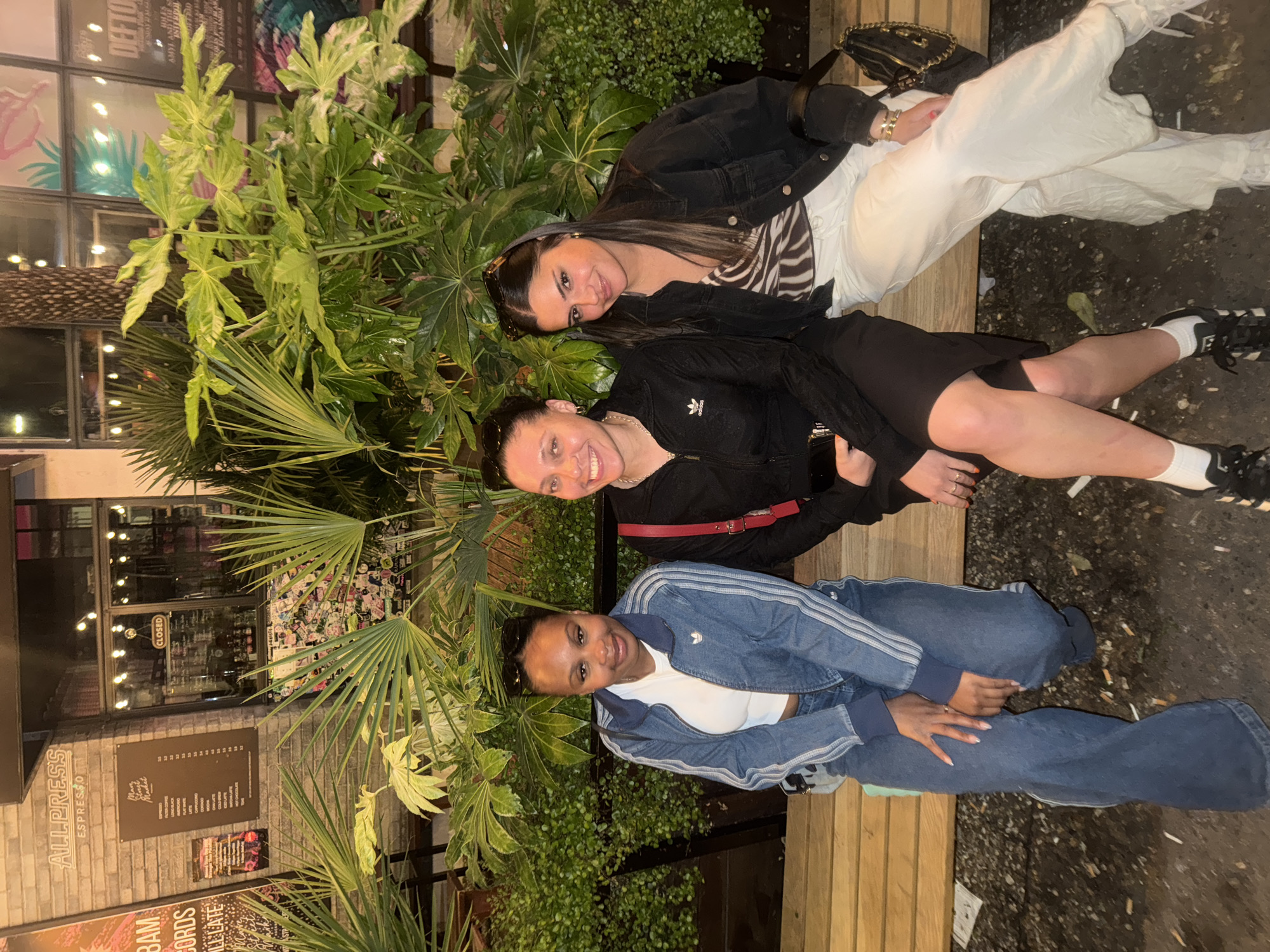 Three women are sitting on a wooden bench outdoors. A building with a menu board reading "Allpress Espresso" is visible in the background. Dense foliage surrounds the bench, partially obscuring the view. The environment appears to be urban, likely in the city of London, United Kingdom.