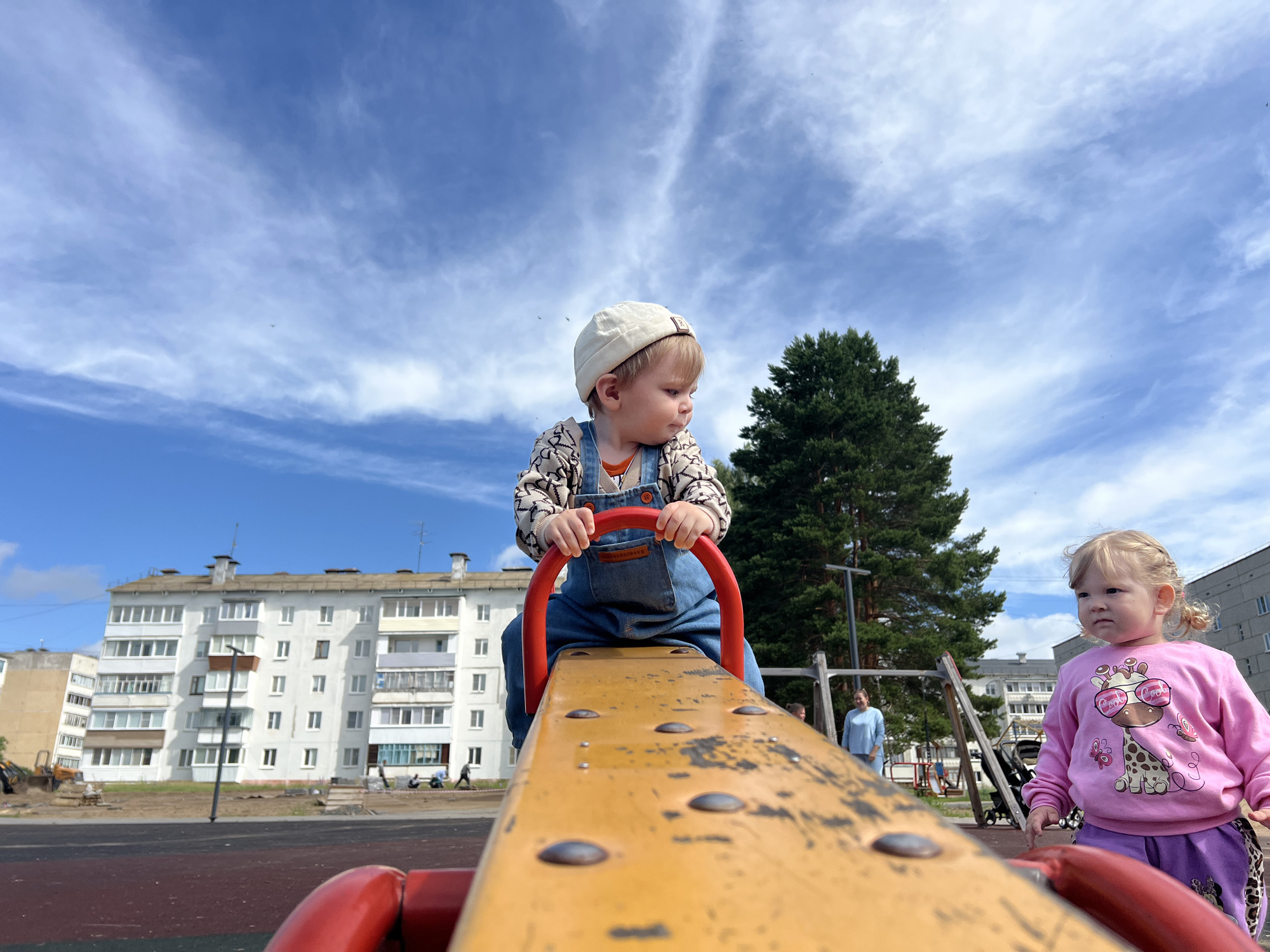 A low-angle, full-shot image captures two young children on a playground in Noviy, Russia, on a sunny day with a partly cloudy sky. In the foreground, a toddler, wearing a white cap, patterned long-sleeved shirt, and blue denim overalls, sits on a yellow seesaw, holding onto the red handles. Their gaze is directed downwards and to the right. To the right of the seesaw, a young girl with blonde pigtails, wearing a pink sweatshirt with a giraffe graphic and purple pants, looks intently towards the left. In the background, a large evergreen tree stands to the right of a multi-story white apartment building. Other children and adults are visible in the distance near playground equipment. The ground of the playground appears to be made of a red and black rubber surface.