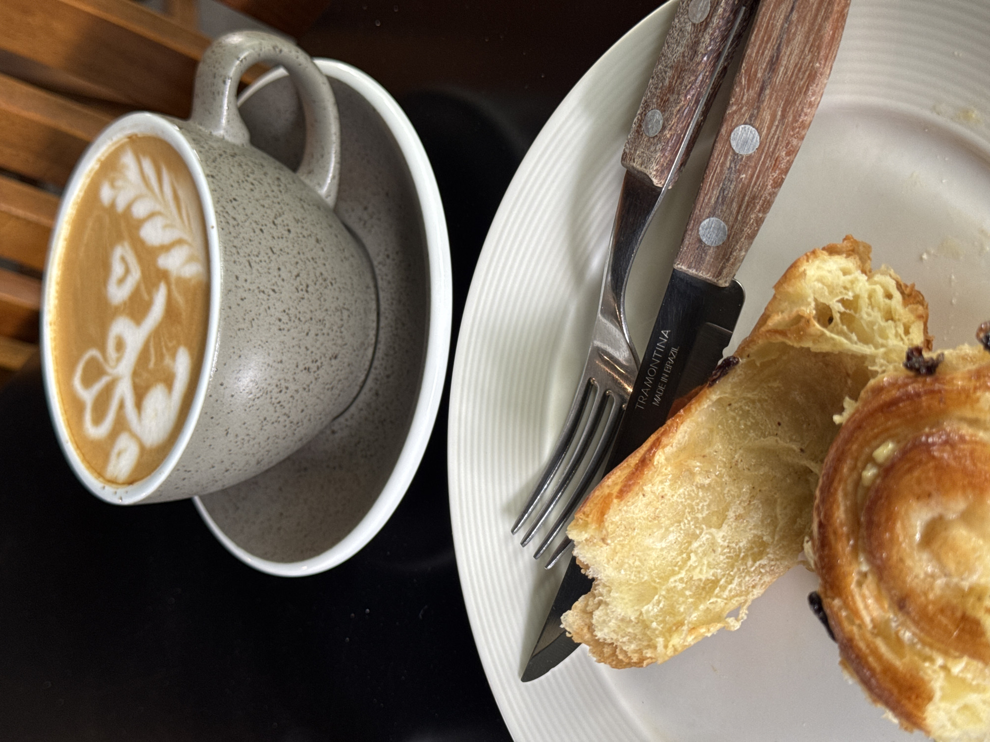 A top-down view shows a speckled gray coffee cup with latte art, placed on a matching saucer to the left. A plate with a partly eaten pain au chocolat and cutlery (fork and knife) are on the right. The knife has the words "TRAMONTINA MADE IN BRAZIL" visible. The scene is on a glossy black surface, possibly a table. Some wooden slats are visible in the top-left corner, suggesting a chair or some other furniture. There is no explicit indication in the image that the location is Kuta Selatan, Indonesia.